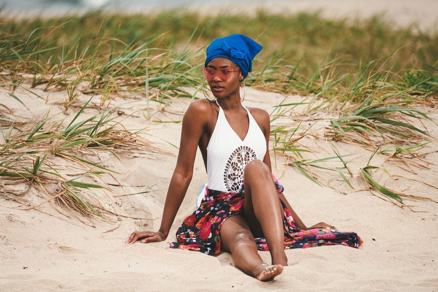 African American woman wearing a white bathing suit and blue head covering as well as pink sunglasses sitting in the sand looking off to the side