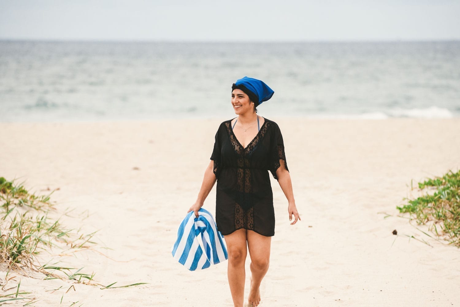 Woman wearing back outfit and blue head covering walking along the beach carrying a blue and white towel smiling looking off to the side