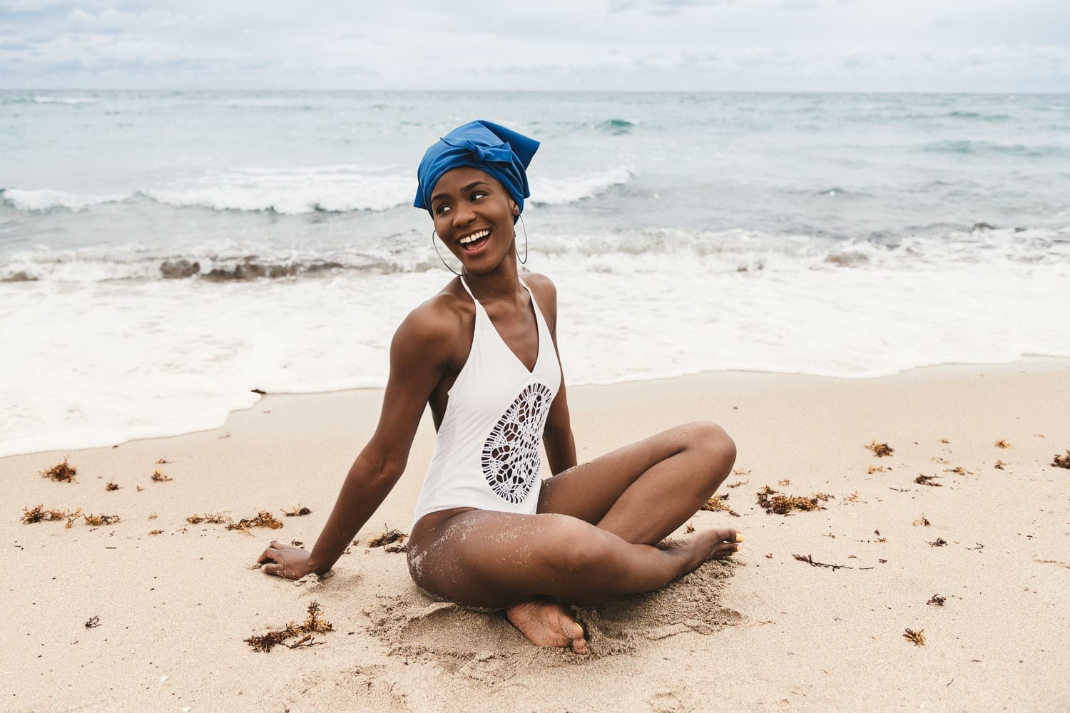 African American woman wearing a white bathing suit and blue head covering sitting in the sand smiling by the water