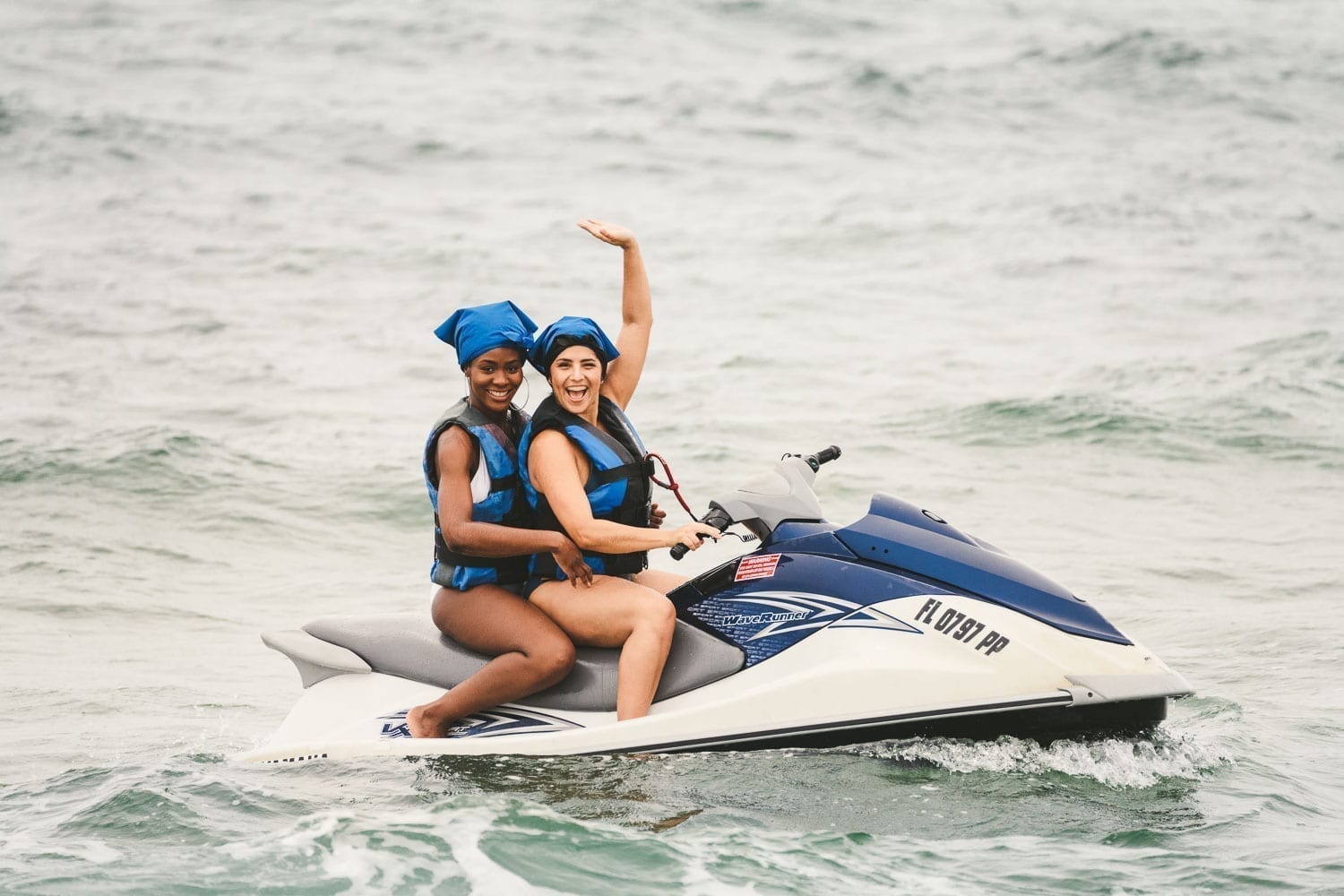 Two women with one African American wearing black and blue life vests and head covering cruising along on a jet ski smiling