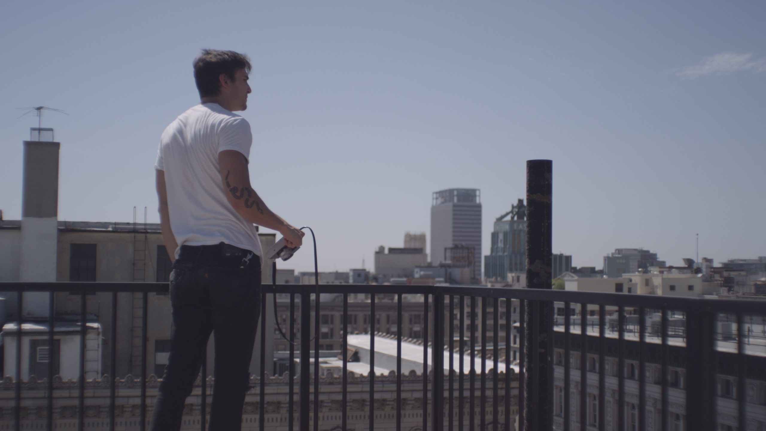 Uncreative Stories Joe Perri Profile View from behind of man with short brown hair wearing a white t shirt looking out over a city holding a camera
