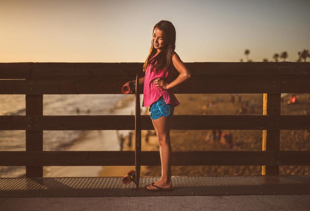 Ivivva Lulu Lemon Side profile of a girl holding a skateboard on a footpath by a beach