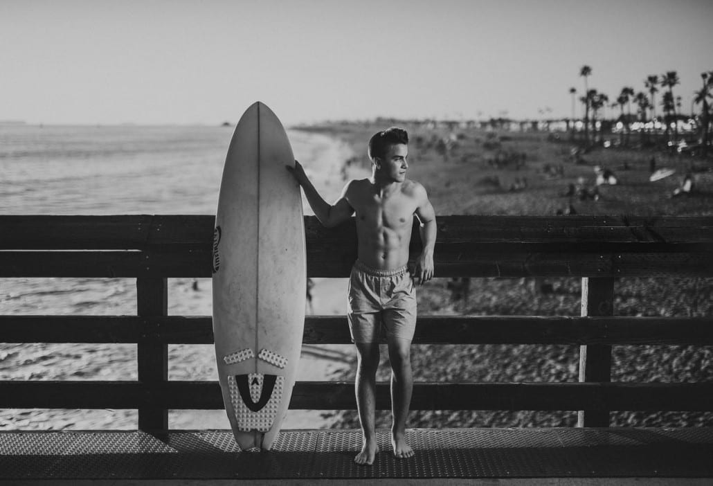 Ivivva Lulu Lemon Black and white of young man holding a surfboard on a footpath by a beach posing for a camera and looking offsides