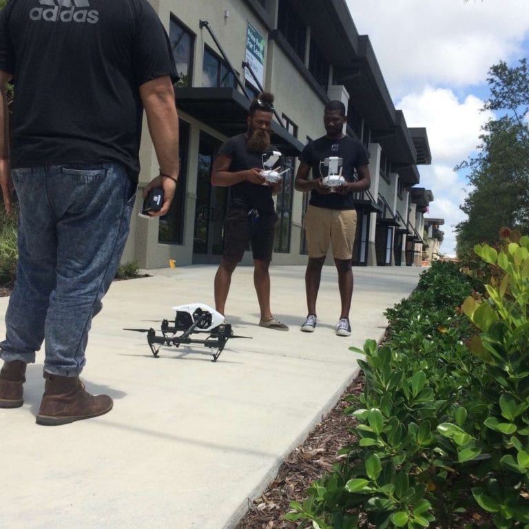 Two men using drone equipment for a drone on the sidewalk while another man looks on