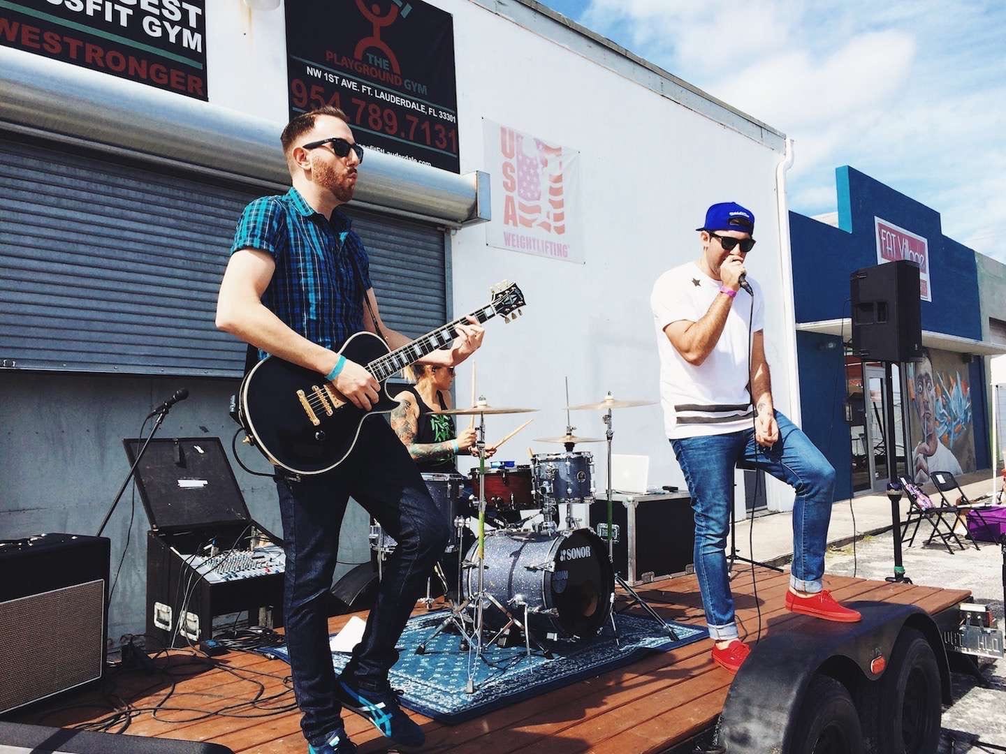 Band consisting of a guitarist, drummer and singer performing on a flatbed trailer in front of storefront at For the Love Music Festival