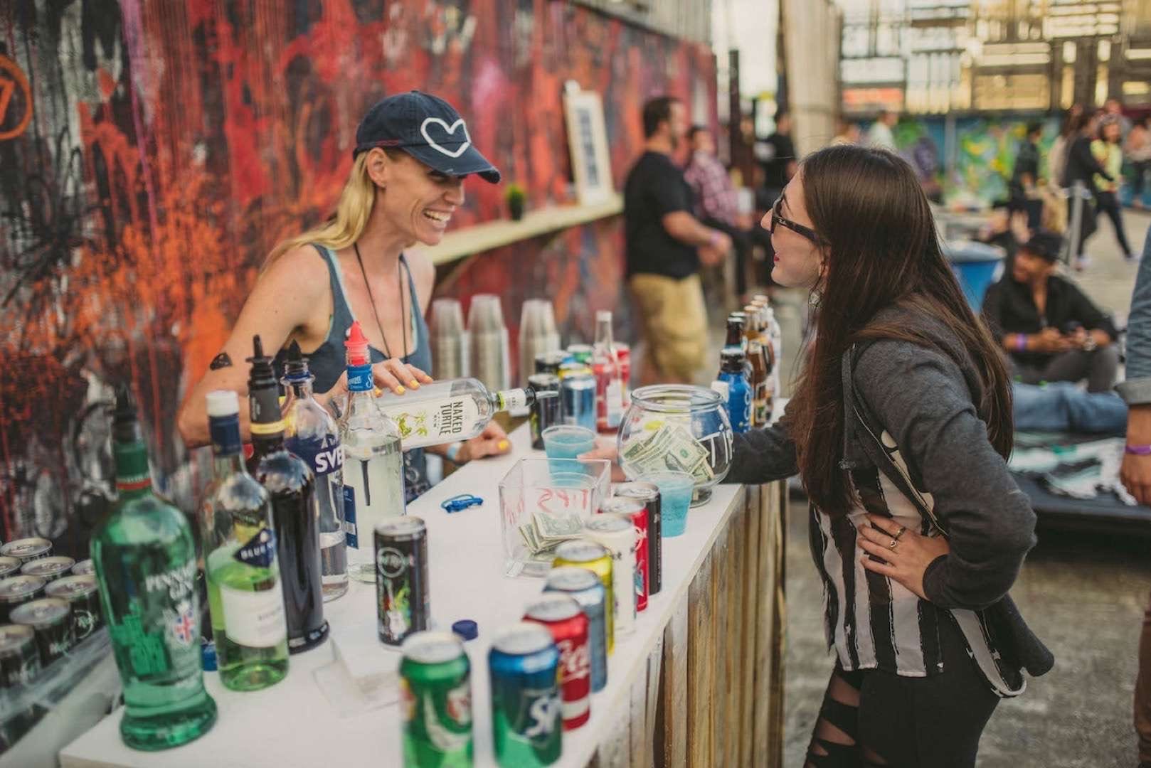 For the Love Music Festival-Smiling bartender serving a drink to a customer at a bar with lots of alcoholic and non-alcoholic drinks on the table