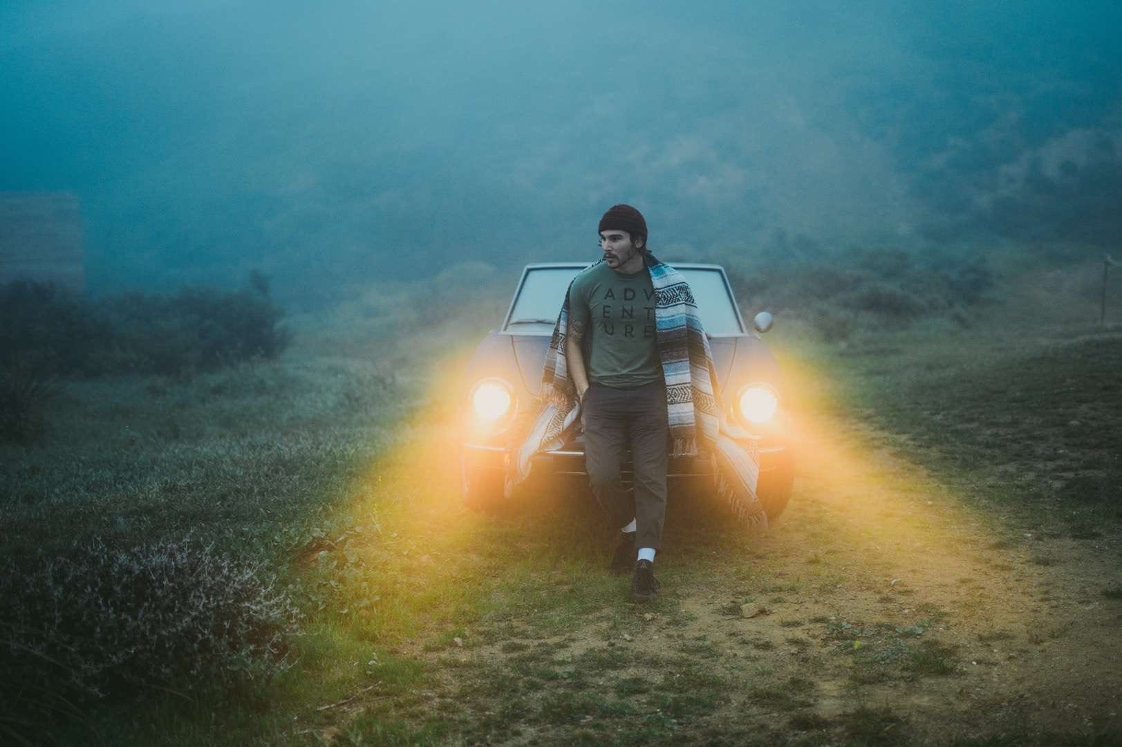 Young man in a clearing at dusk with Indian style blanket draped over shoulders leaning back against the hood of a car with blazing headlights looking off to the distance.