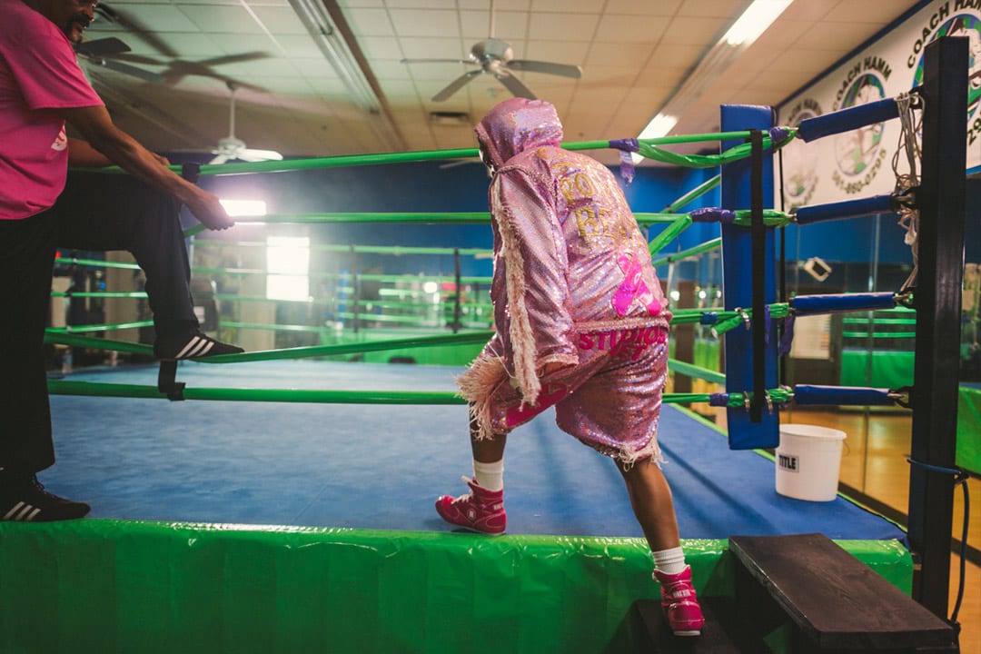 Gold Coast Boxer Woman in pink outfit climbing onto the blue boxing mat with light green boundaries. Her training is climbing in nearby. There is a white bucket on the mat nearby.