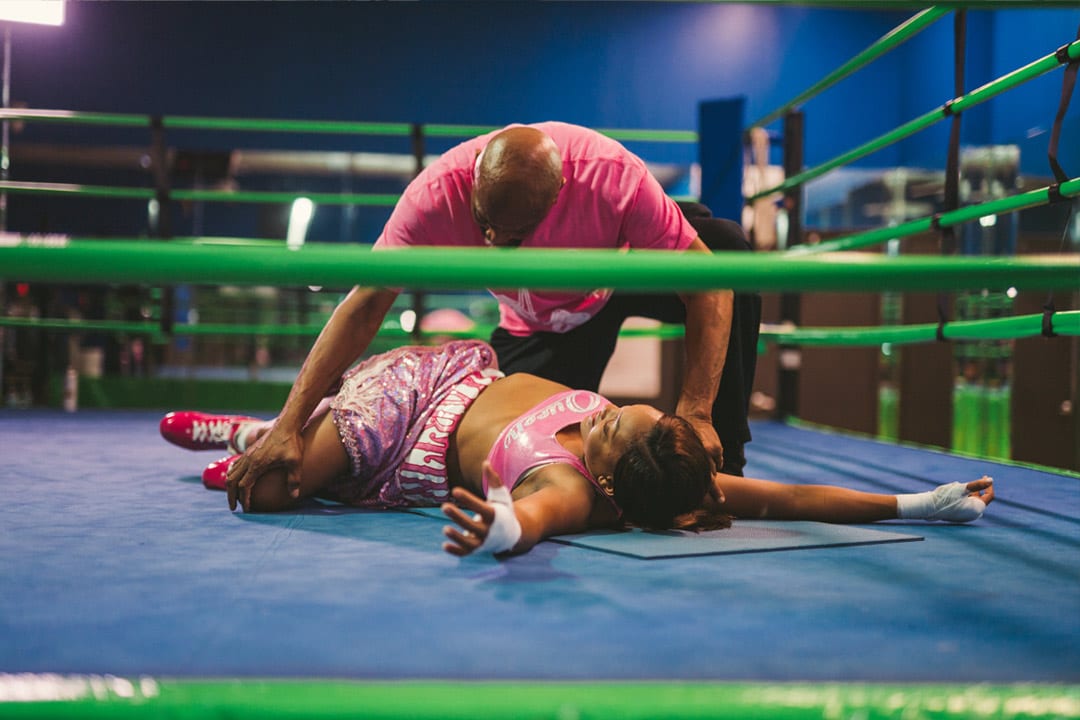 Female boxer in pink outfit lying down on dark blue mat with light green boundaries. She is wearing bright red shoes. Her trainer is helping with leg related stretching.