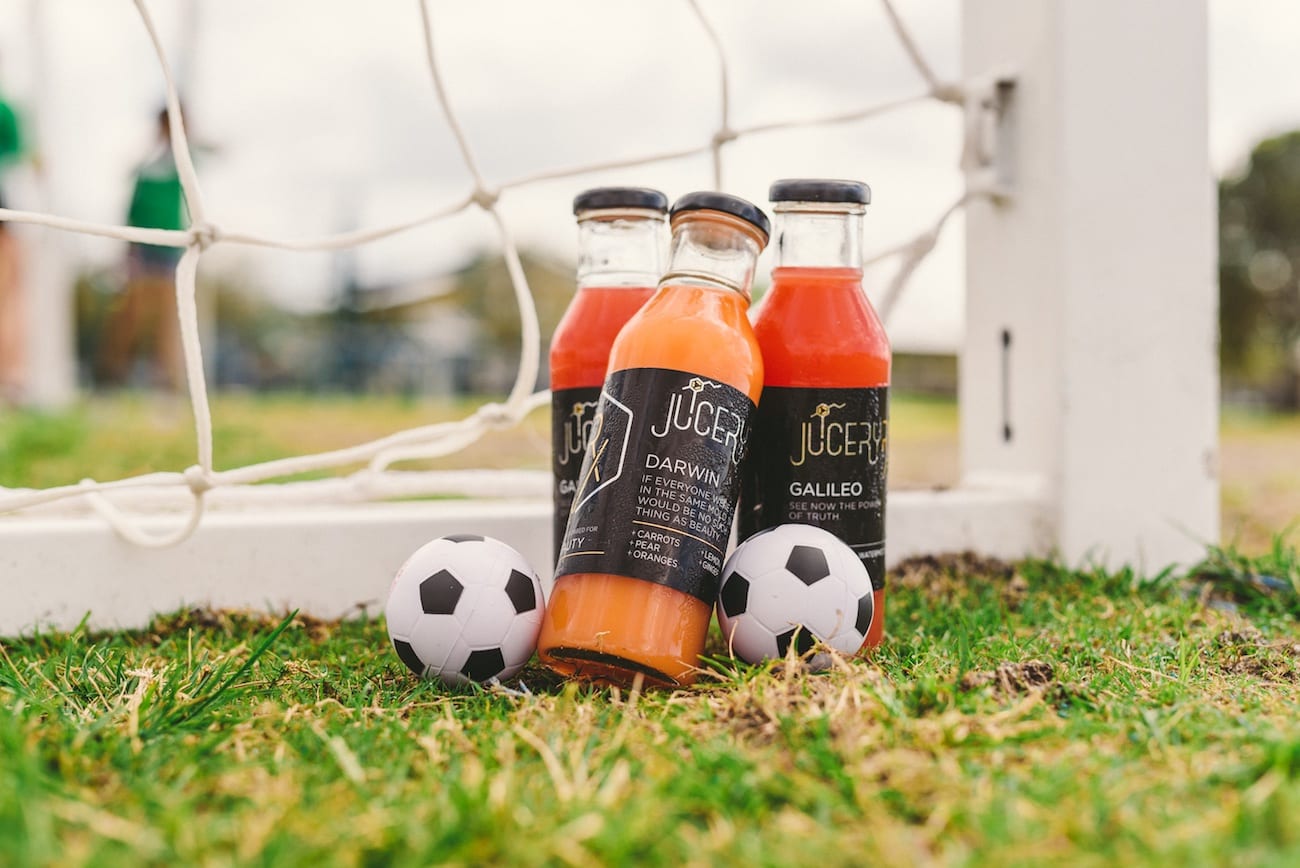 Three varieties of glass bottles of juice on display by a soccer net with tiny soccer balls