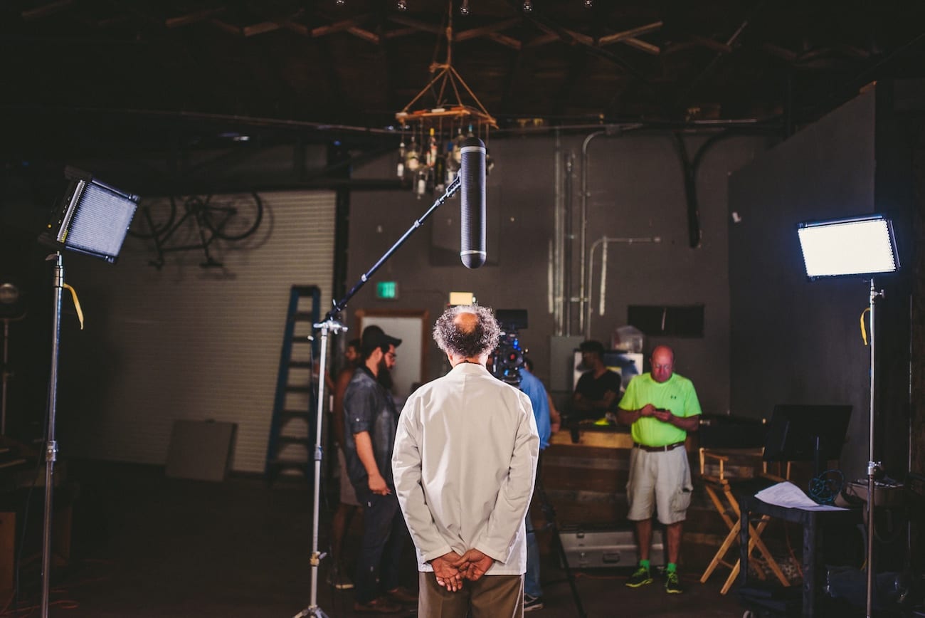 View from behind of a man in a white lab coat waiting for to start his GhostBed pitch with filming crew and equipment in the background