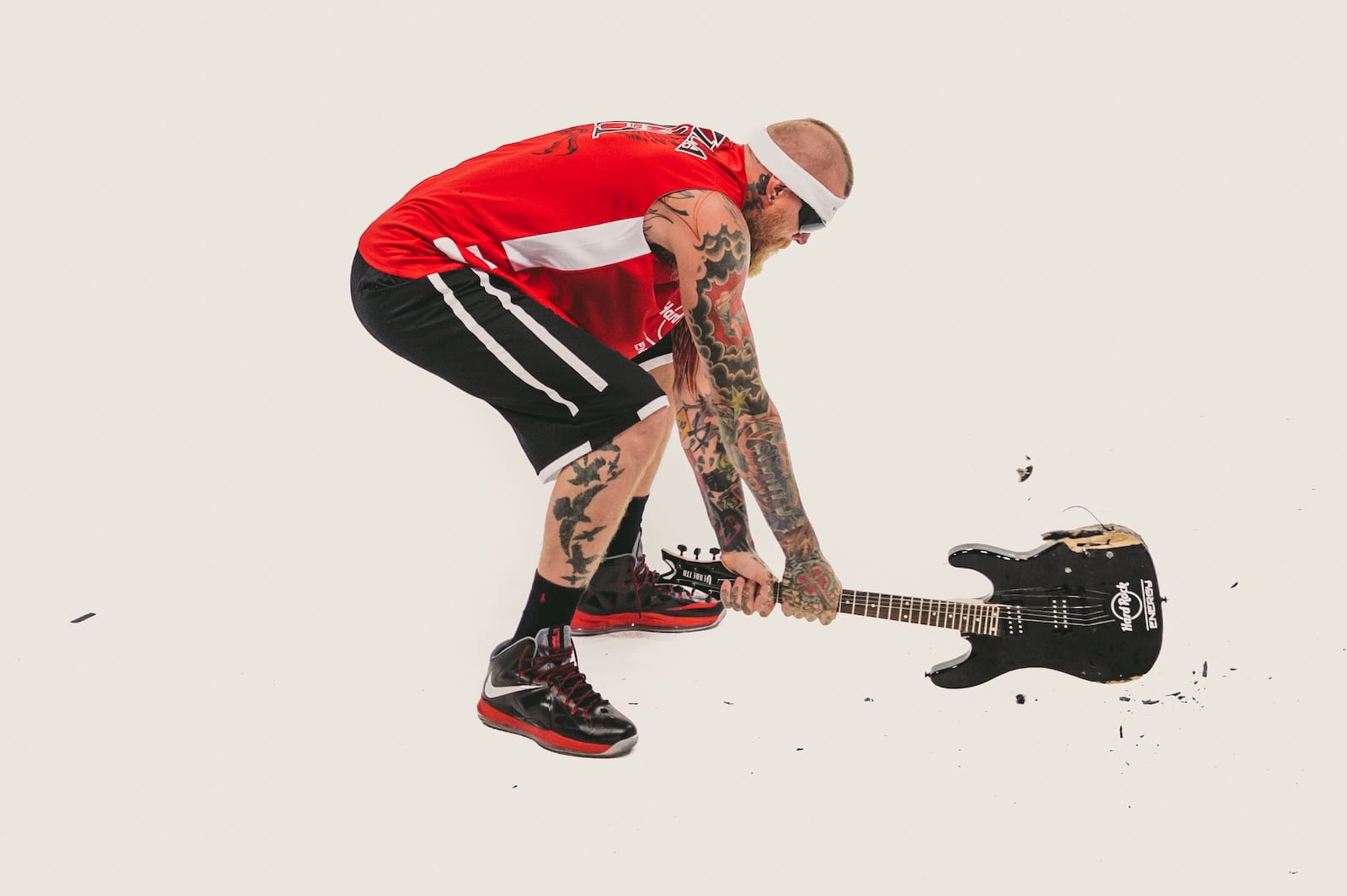 Side profile of a bearded tattooed man wearing sunglasses slamming a guitar on the floor wearing red Hard Rock Energy jersey with black shorts