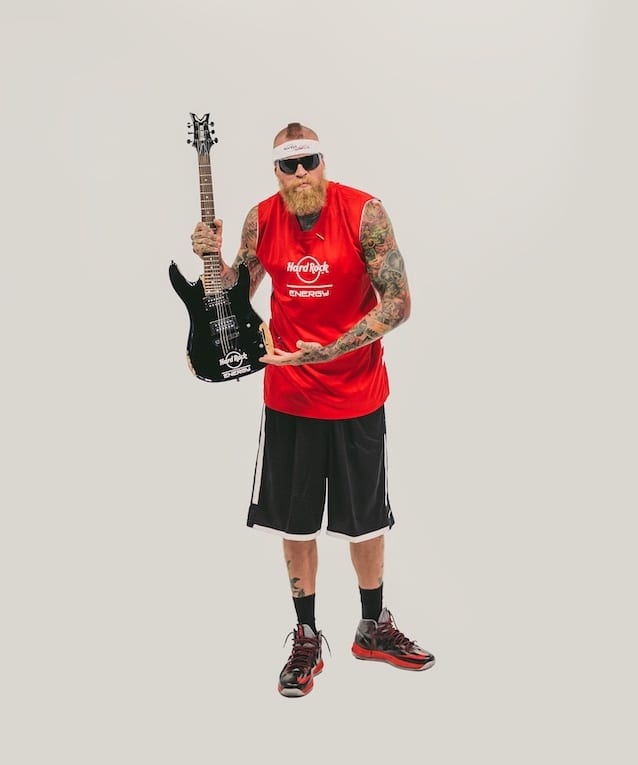 Bearded tattooed man wearing a red Hard Rock Energy jersey and black shorts holding a guitar