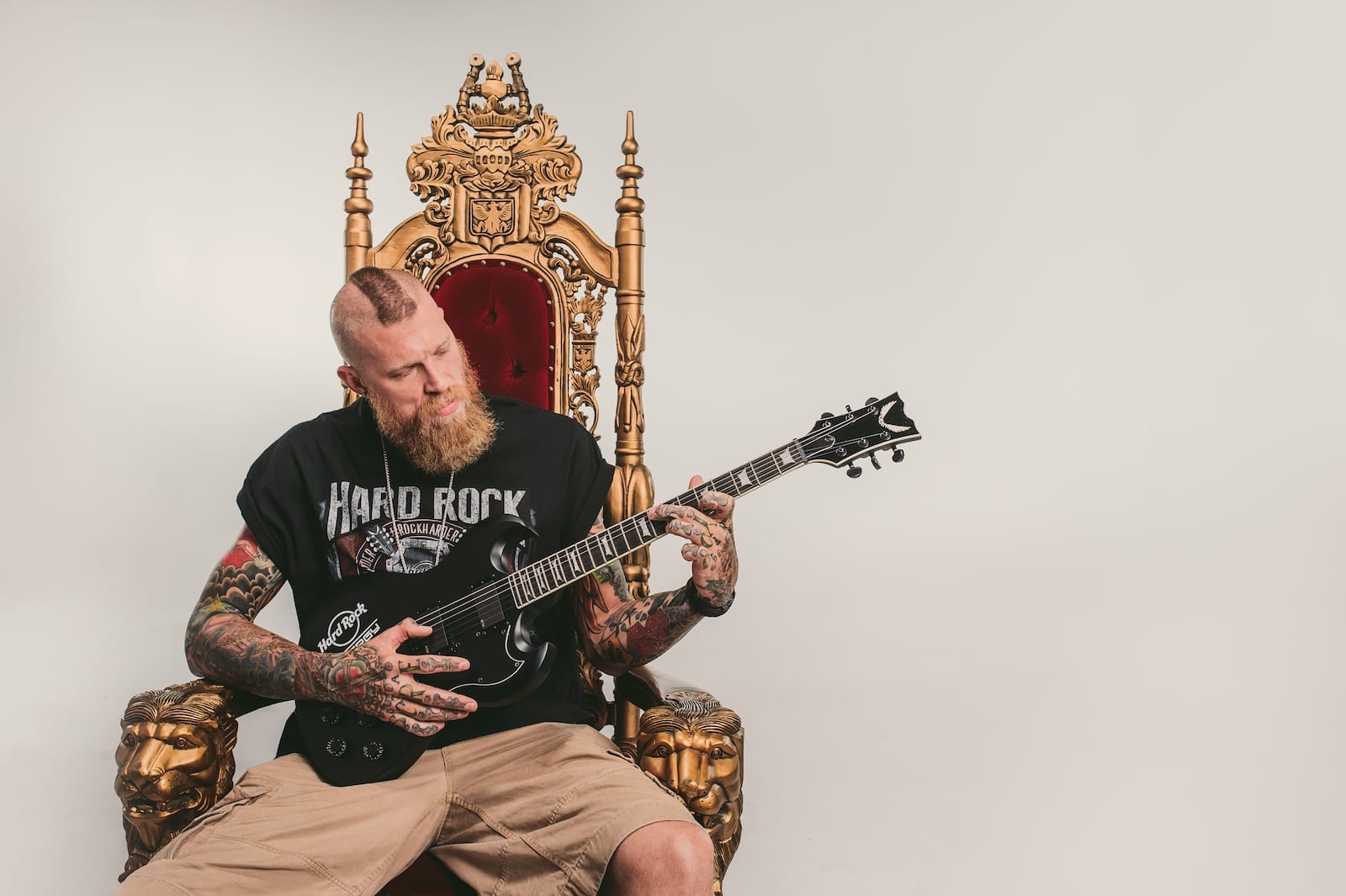 Bearded tattooed man playing a guitar on a throne posing for camera wearing a black Hard Rock tshirt