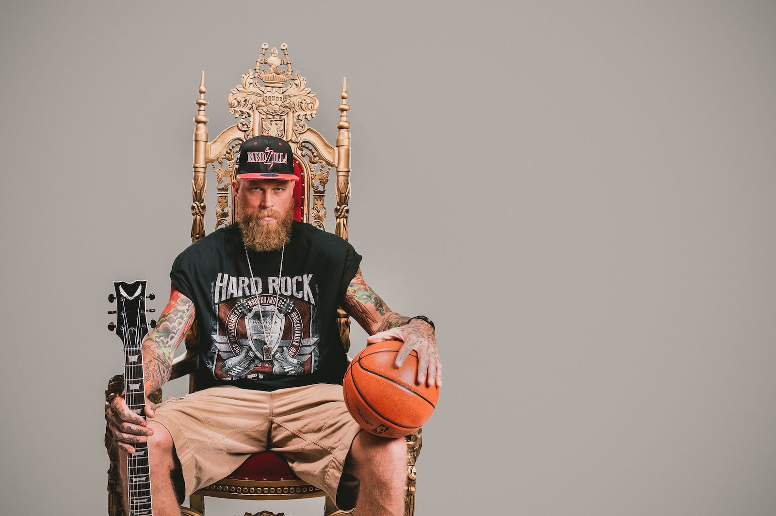 Bearded tattooed man holding guitar in one hand and a basketball in the other sitting on a throne posing for camera wearing a black Hard Rock tshirt
