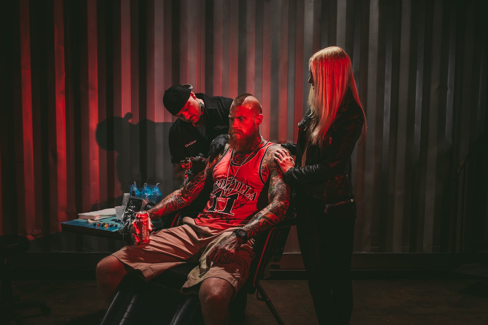 Bearded tattooed man holding Hard Rock Energy drinks with tattoo artist working on a tattoo with an assistant standing nearby