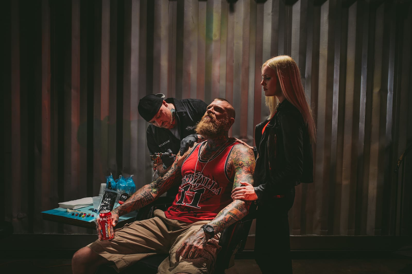 Bearded tattooed man holding Hard Rock Energy drinks with tattoo artist working on a tattoo with an assistant standing nearby