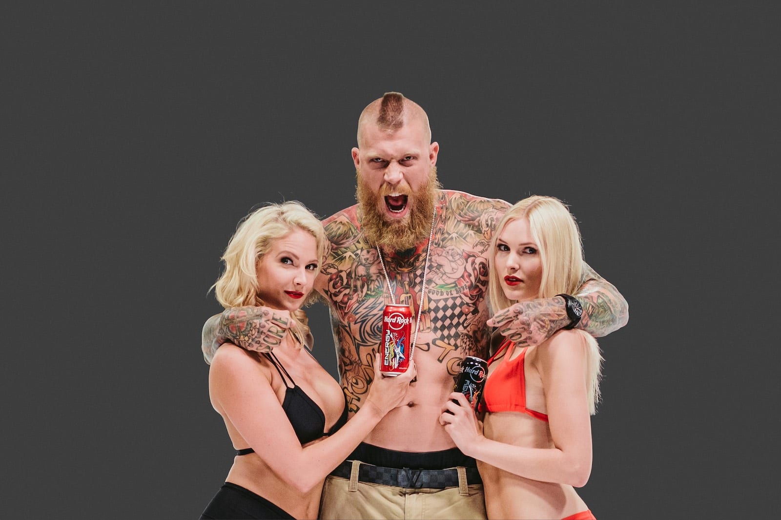 Bearded tattooed man holding two women holding Hard Rock Energy drinks next to him posing for camera