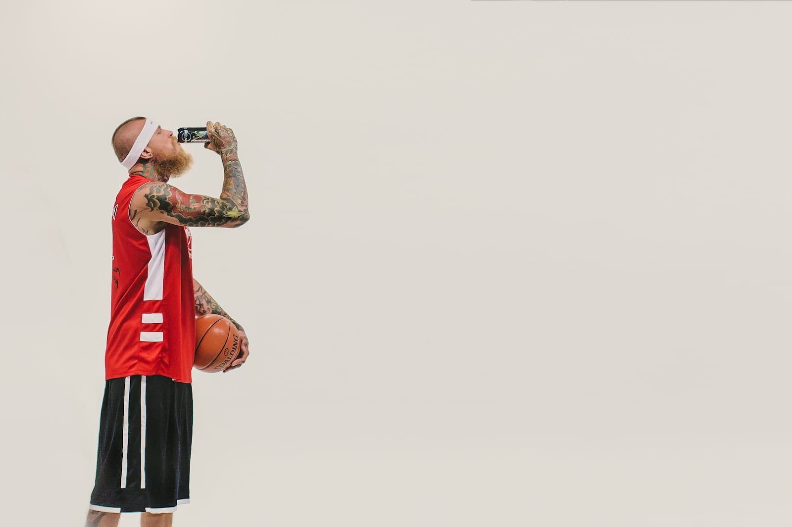 Side profile of a bearded tattooed man drinking from can of Hard Rock Energy drink wearing red Hard Rock Energy jersey with black shorts holding a basketball