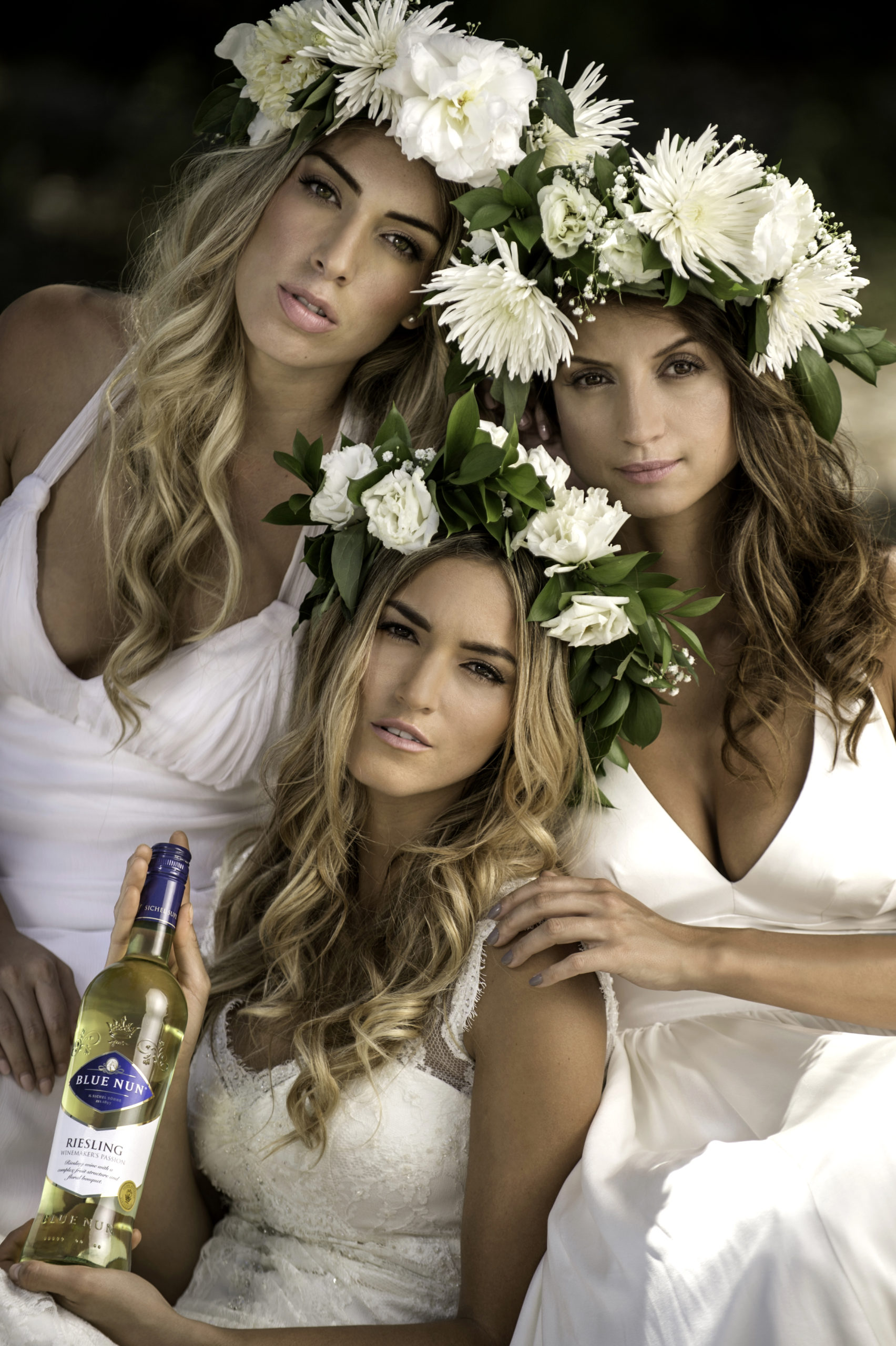 Three women wearing flower headdresses posing for the camera wearing white dressing gowns with one holding a bottle of Riesling wine