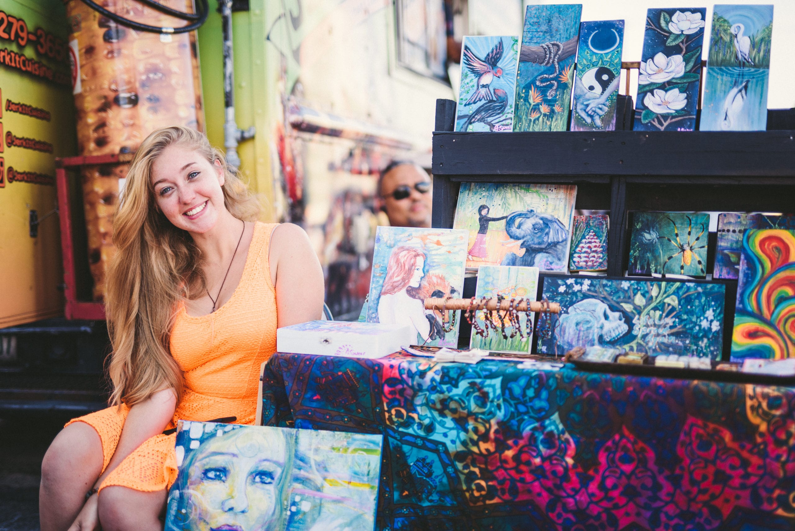 Woman with long blond hair wearing orange dress posing next to artwork
