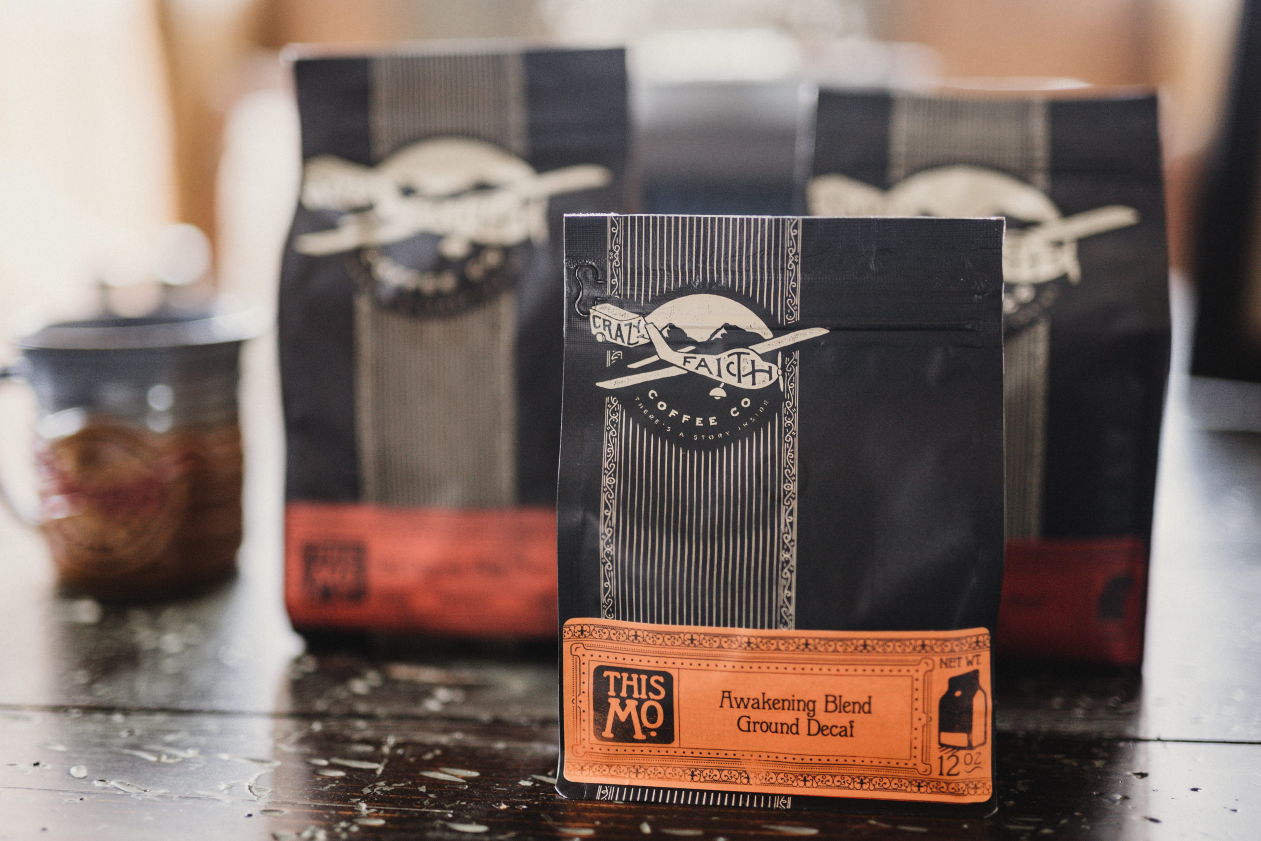 Packages of Crazy Faith coffee grounds with Awakening Blend Ground Decal blend in the foreground with a brown and blue coffee cup nearby