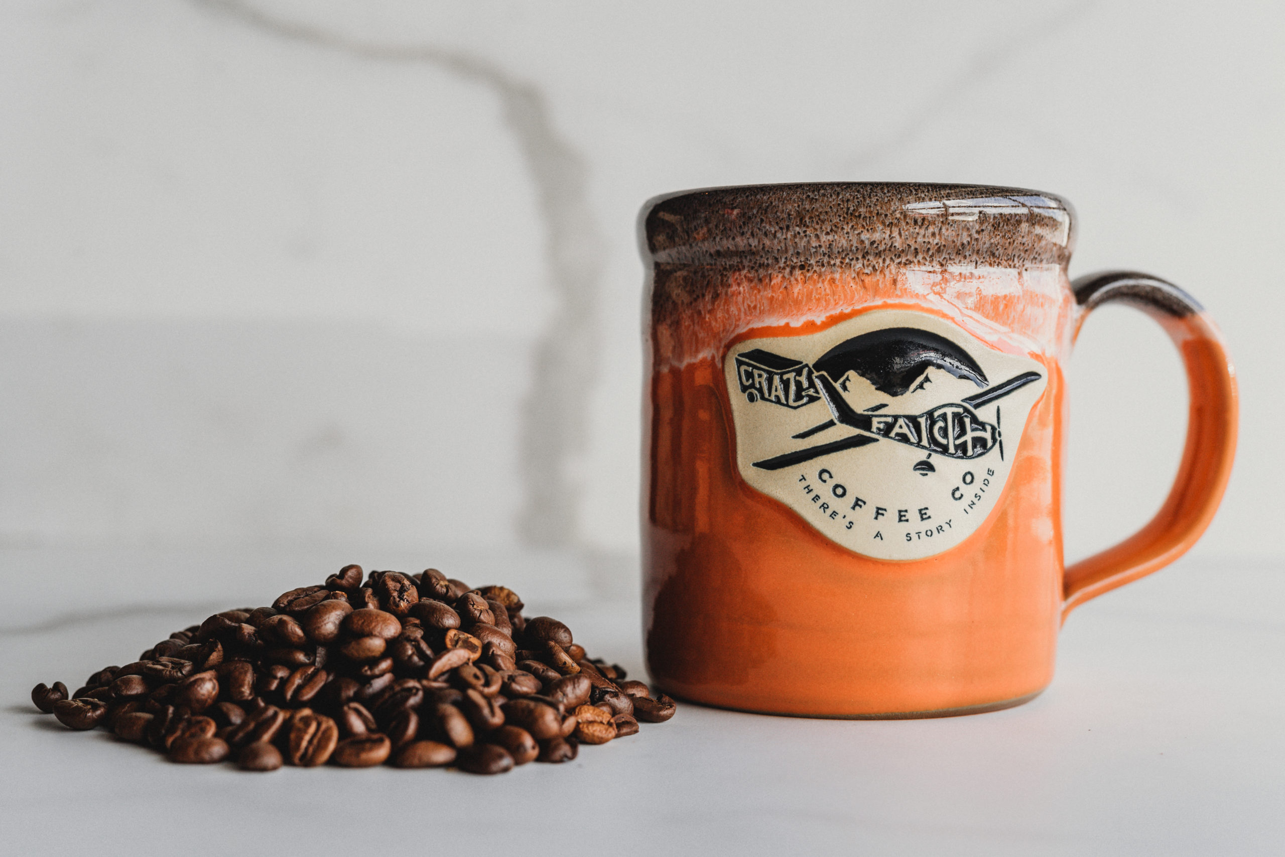 Orange and brown Crazy Faith coffee cup next to coffee beans
