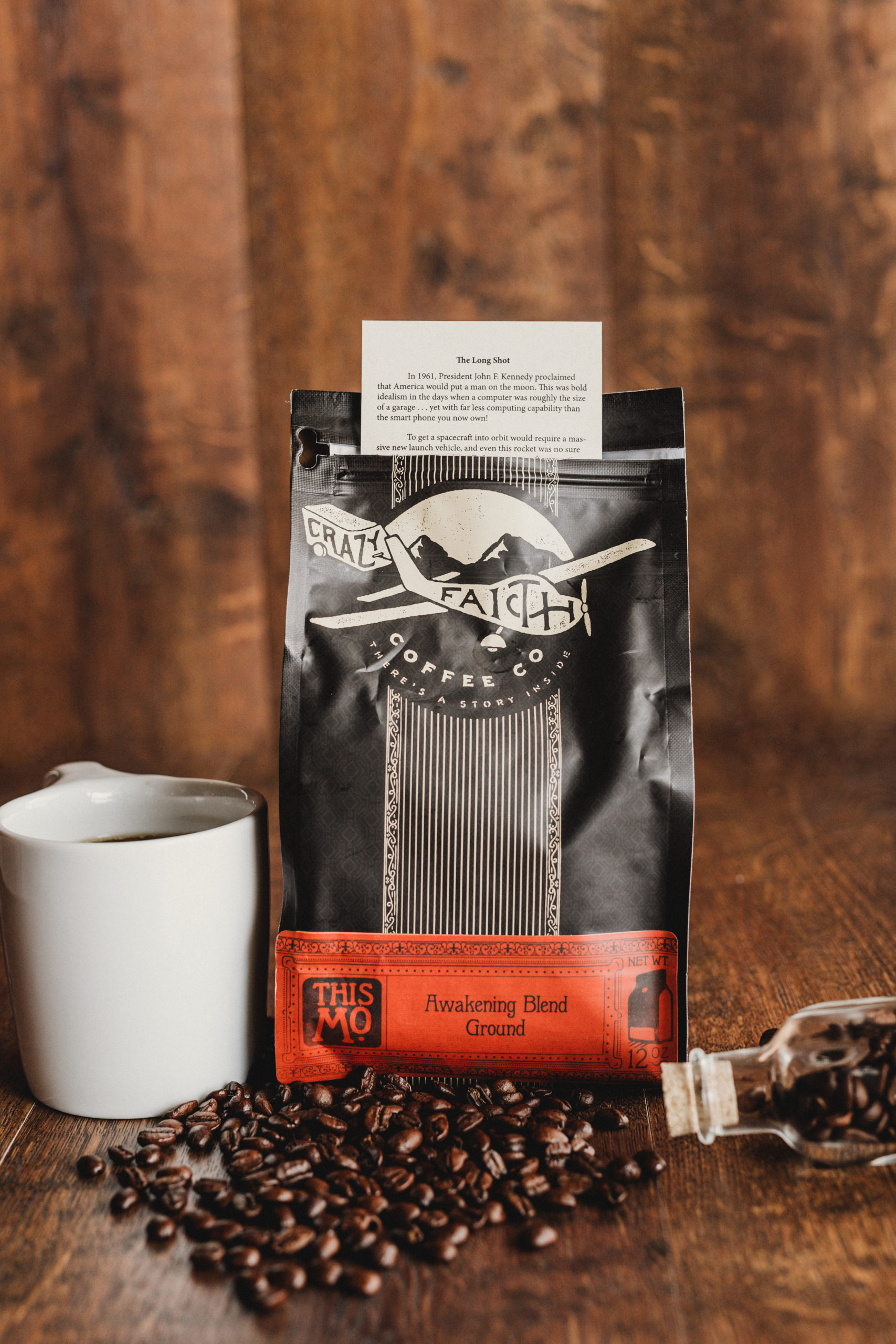 Package of Awakening Blend Ground coffee beans along vial and coffee beans on the wooden table along with white coffee cup with coffee