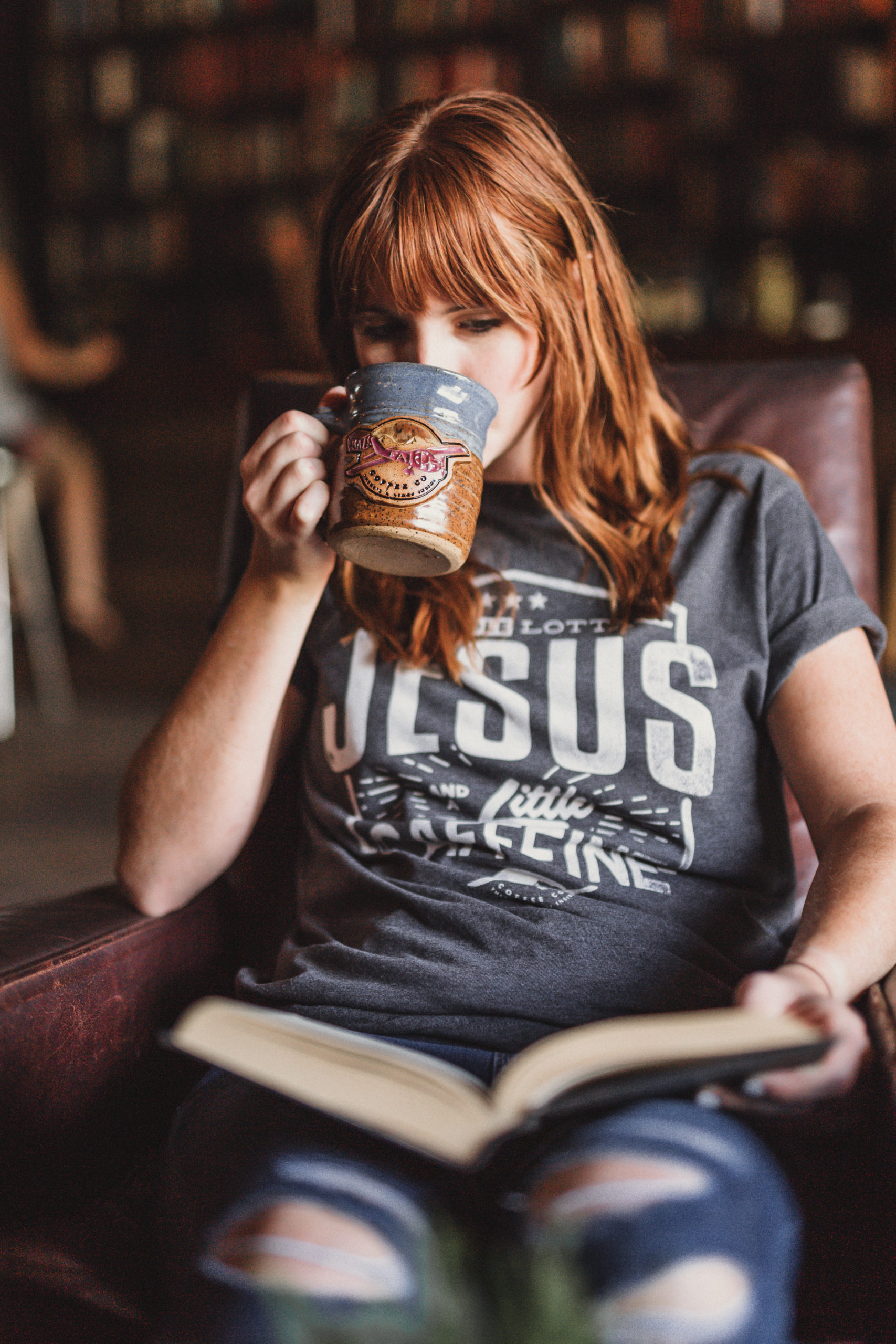 Woman wearing a gray t shirt that says A Whole Lotta Jesus and a Little Caffeine from Crazy Faith drinking some coffee from a brown and gray coffee cup and reading a book