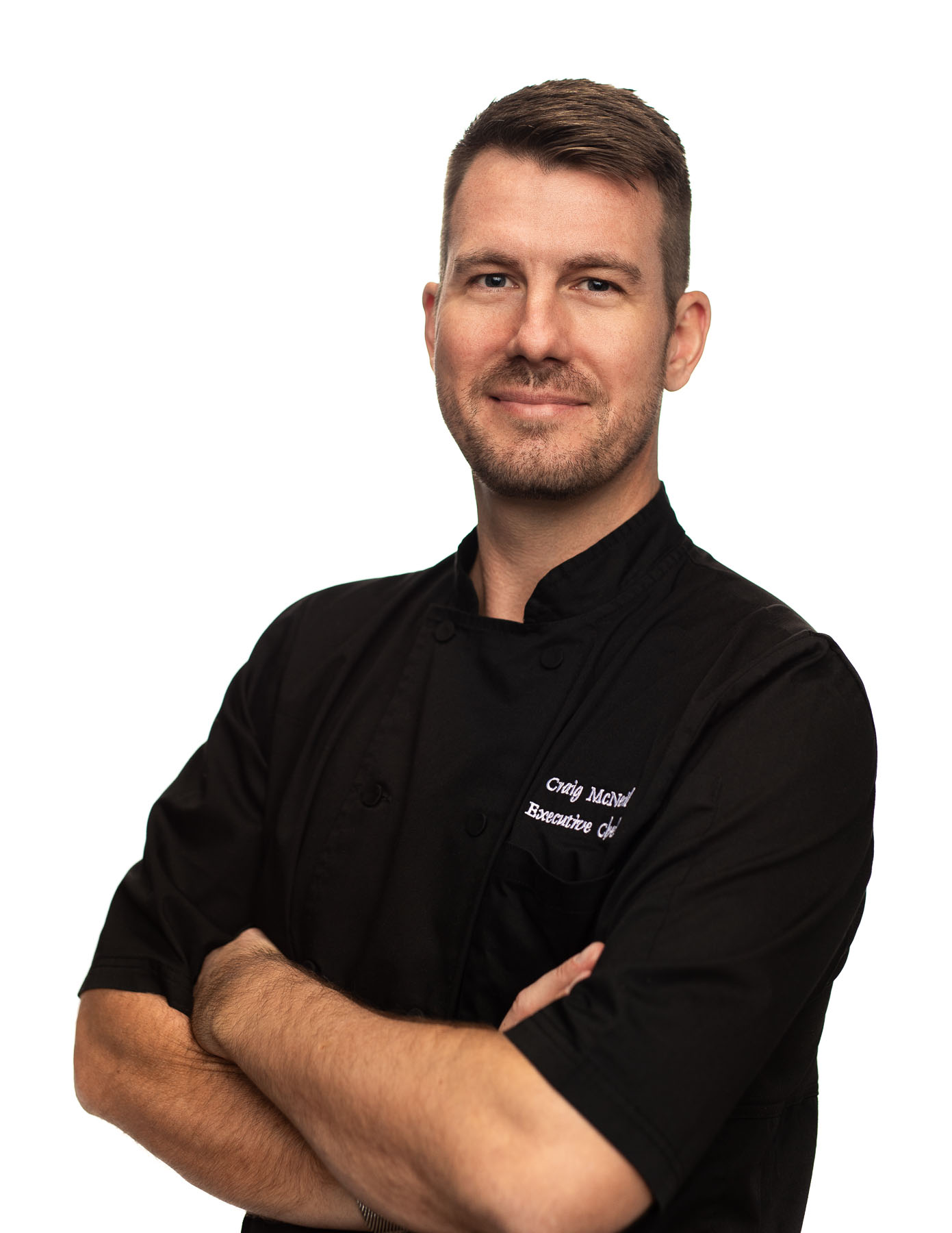 Headshot of man with short hair wearing a black tshirt with label Craig McNeil Executive Chef posing and smiling for the camera with arms crossed