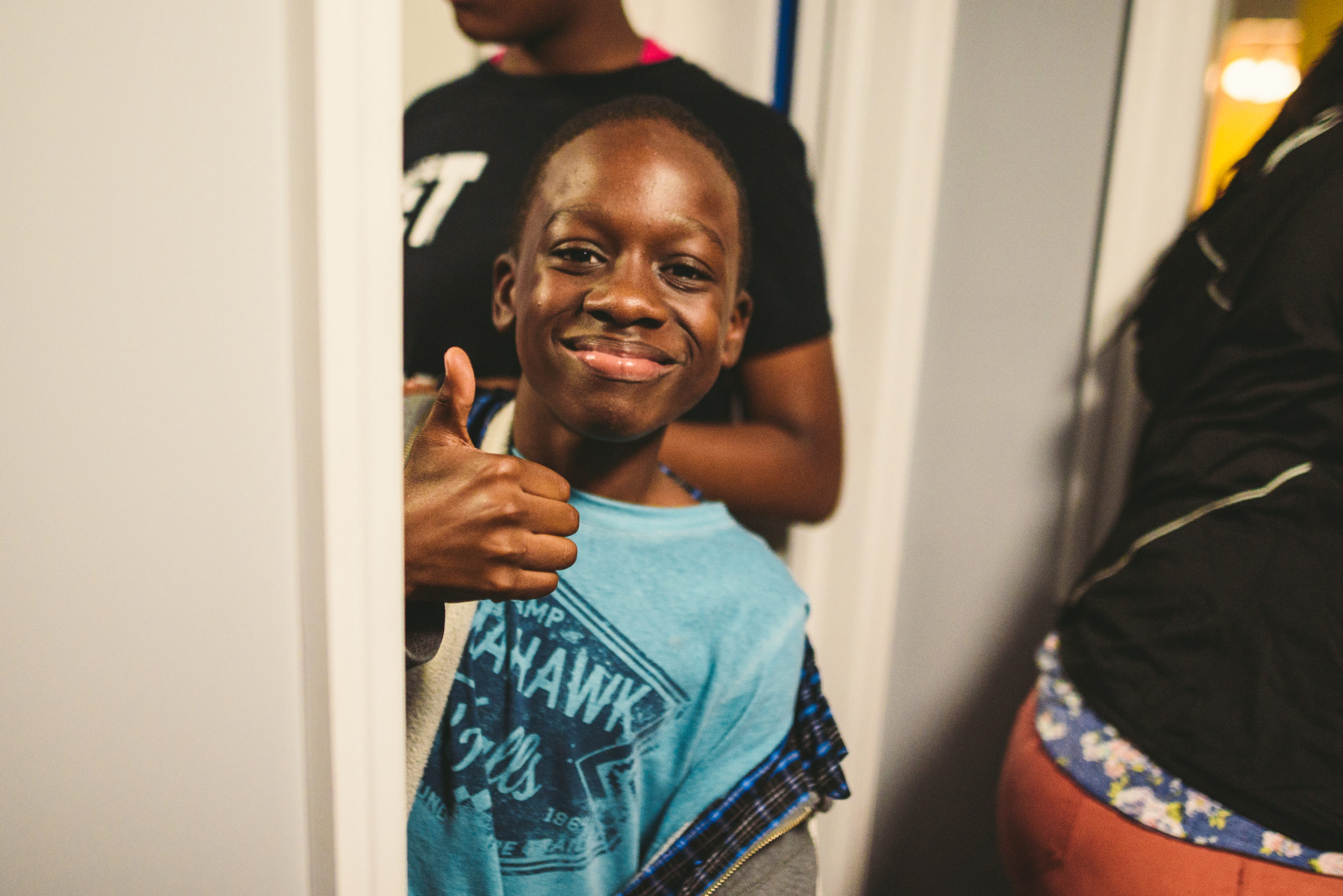 IU CI Studios Handy Nonprofit African American boy wearing a light blue shirt with logo giving a thumbs up and smiling surrounded by people