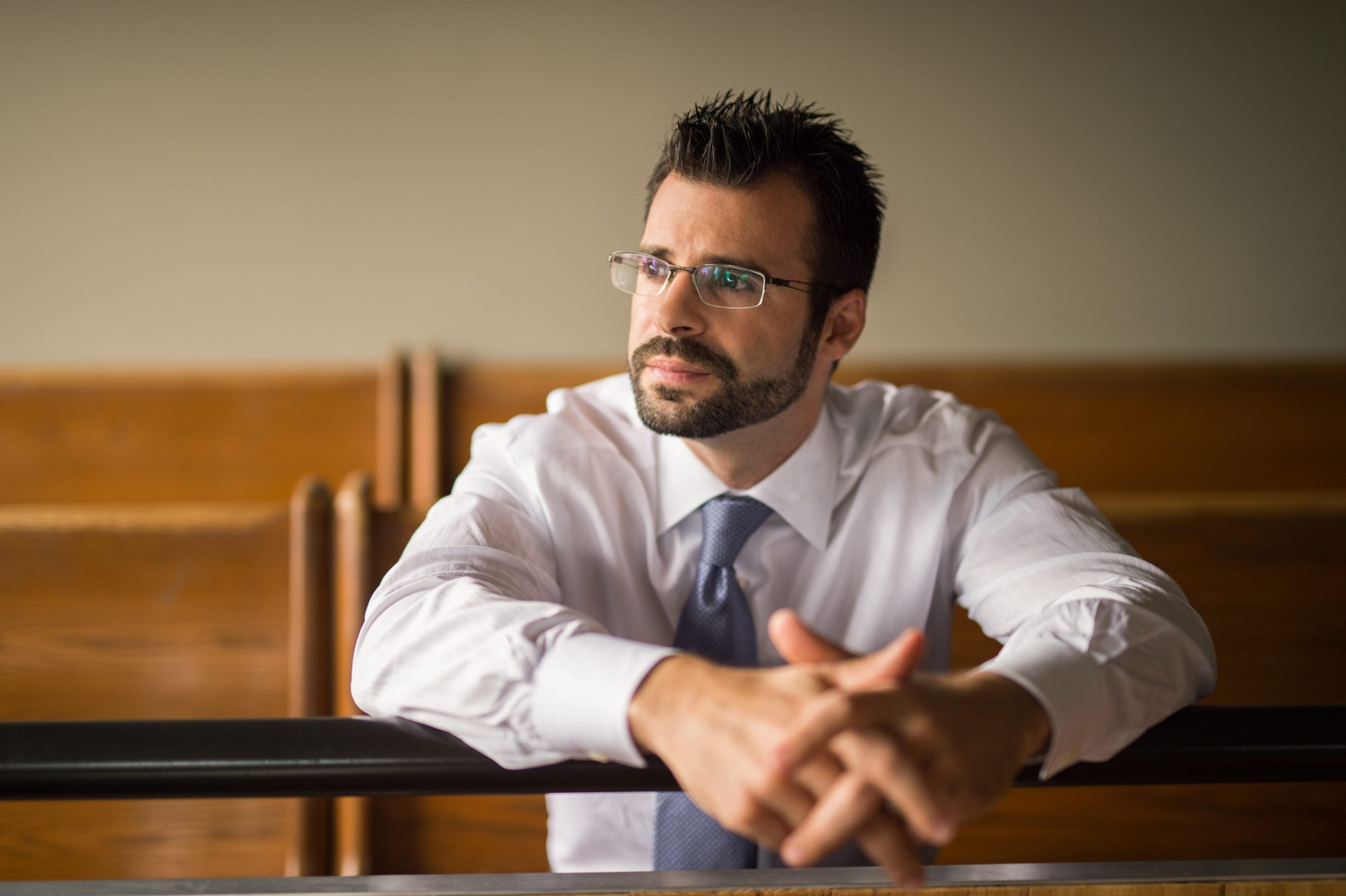 Kelley & Uustal Trial Attorneys Headshot of a male attorney with a beard wearing a white shirt, blue tie and glasses leaning on the railing in a courtroom looking off camera