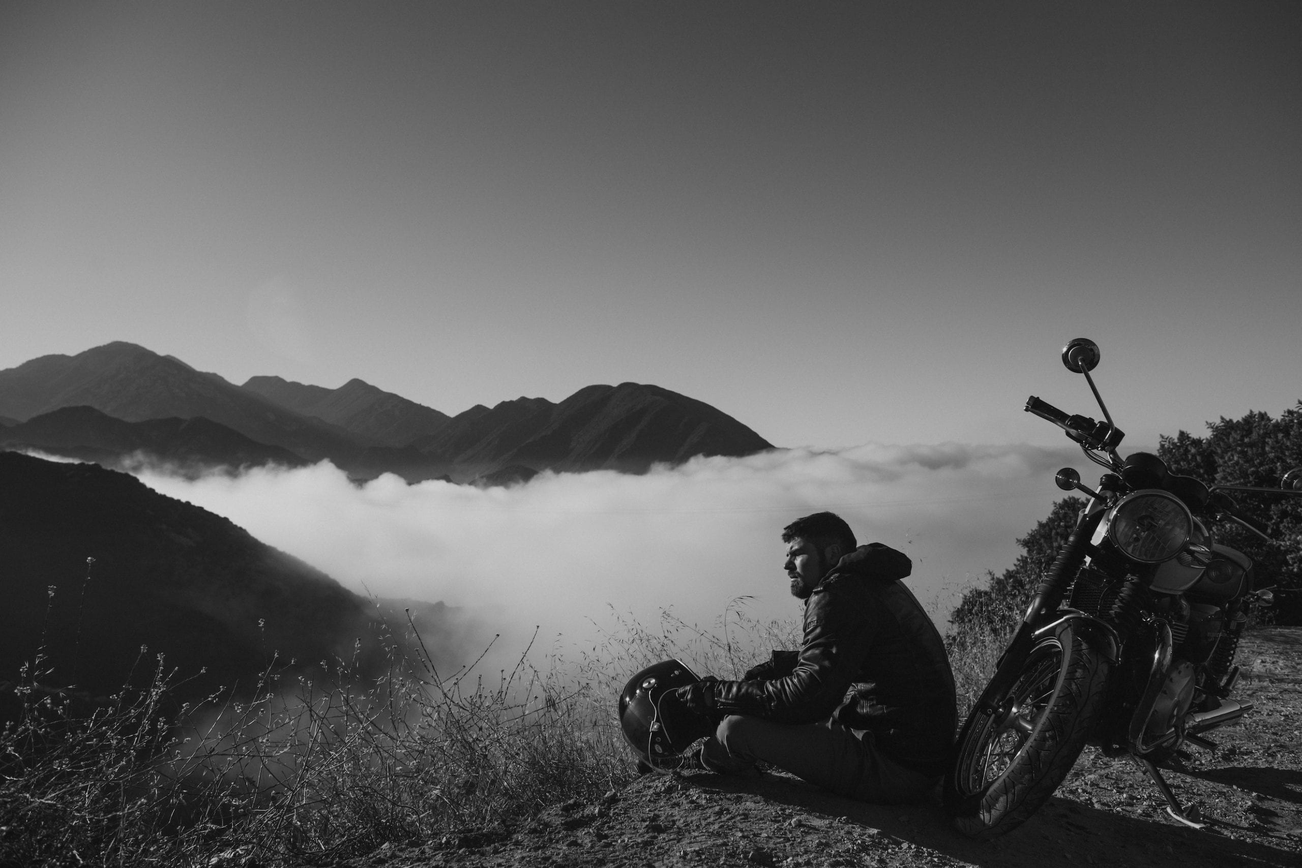 Black and white photo of a motorcycle rider resting above the clouds overlooking a mountain range.