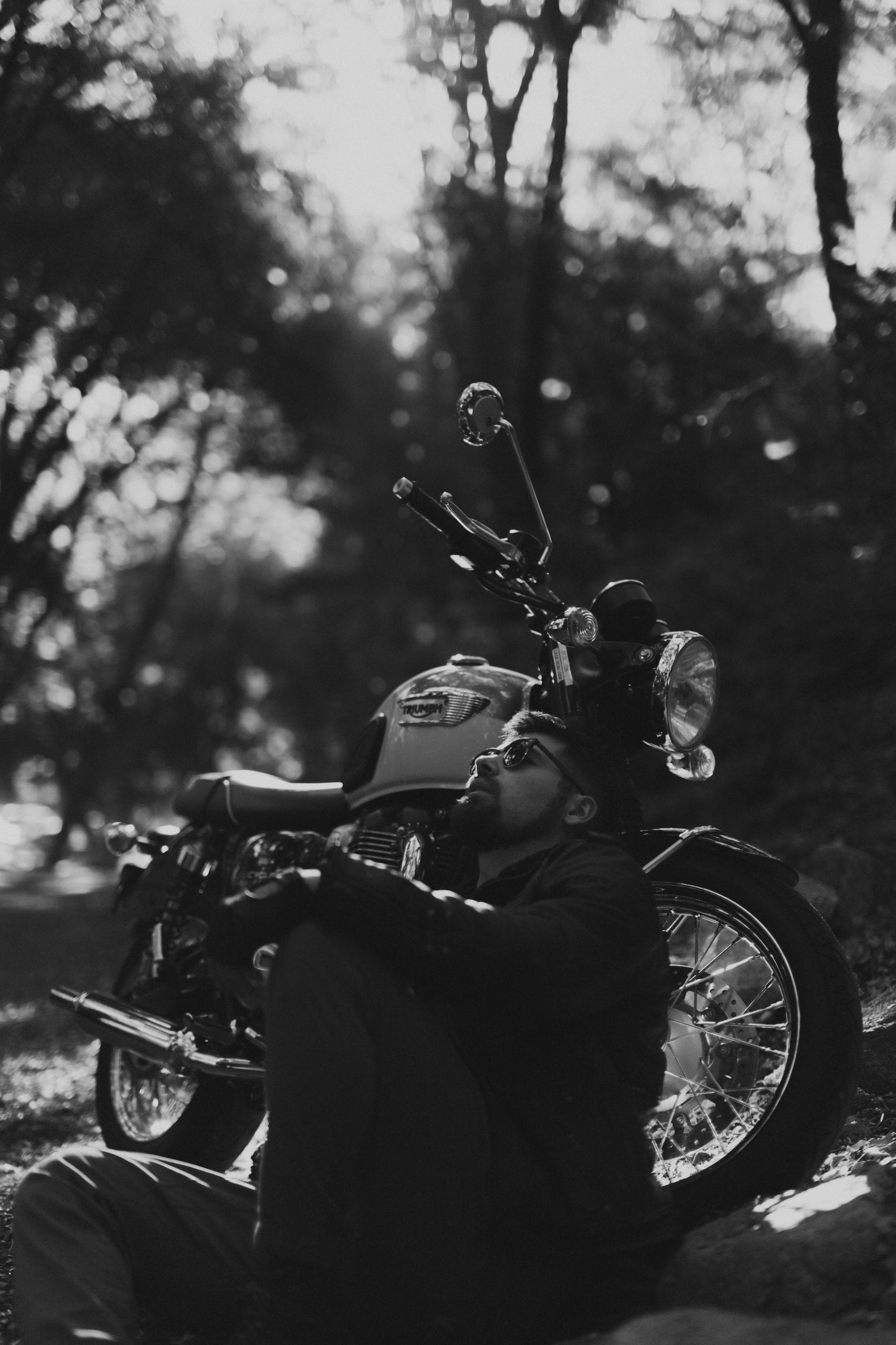 Black and white of motorcycle rider wearing shades relaxing sitting near the motorcycle posing for the camera and looking up at the sky
