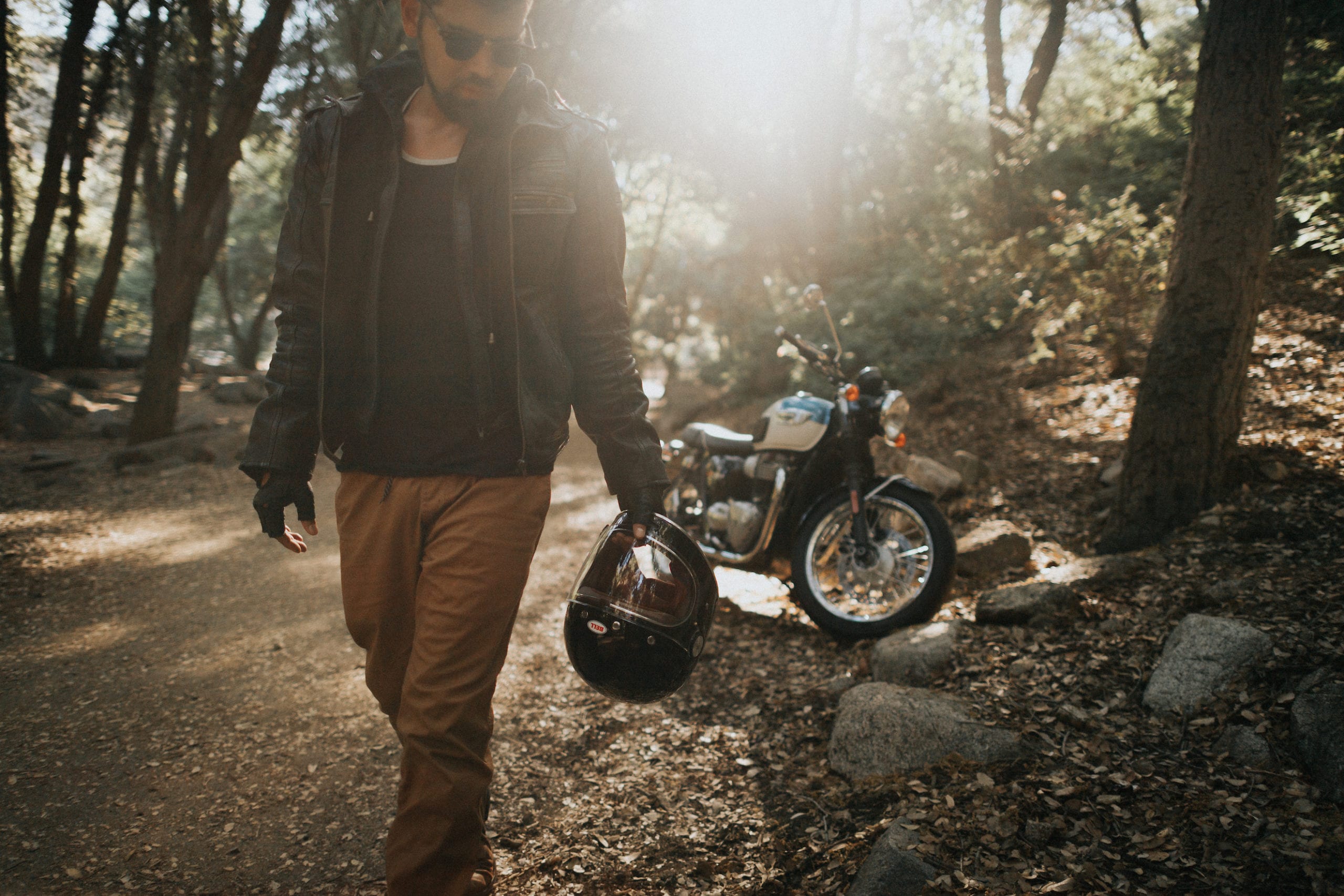 Motorcycle rider wearing shades and leather gear poses for the camera in the middle of the forest holding his Bell helmet