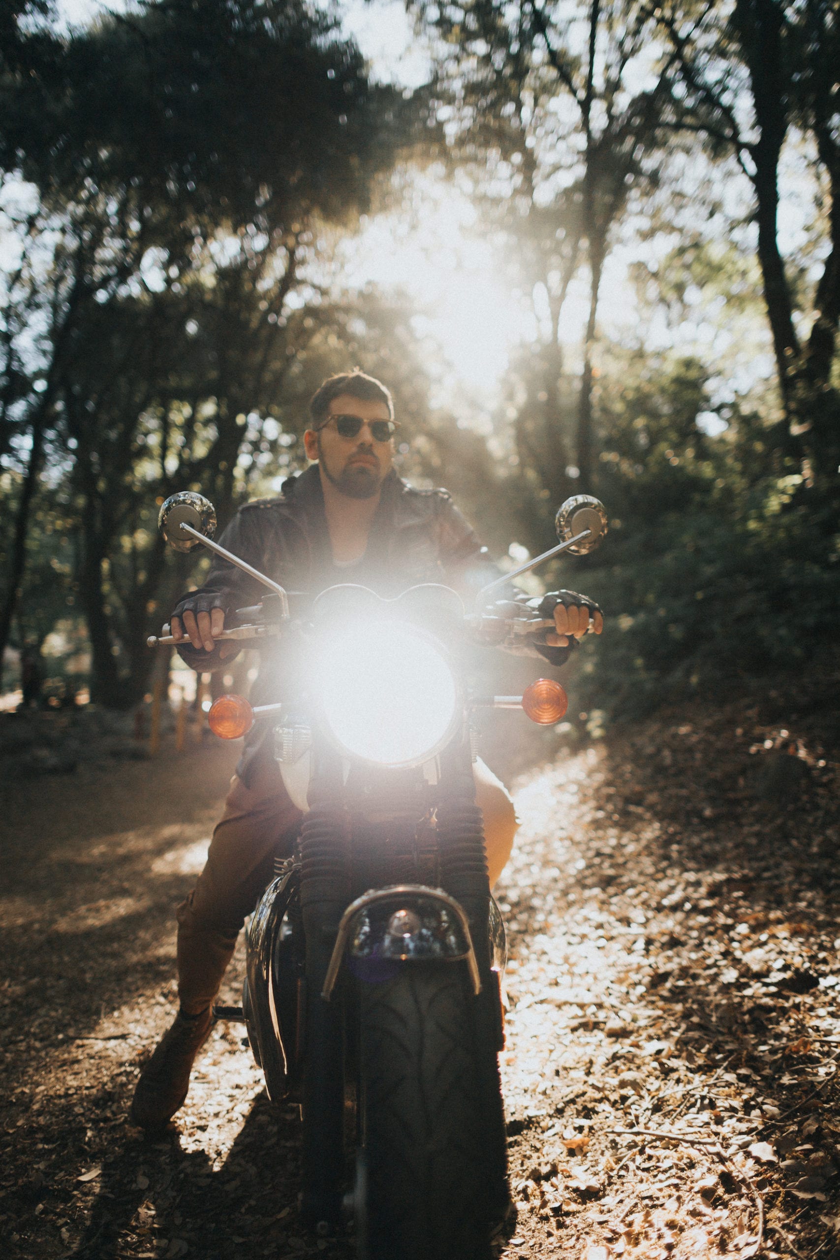 Motorcycle rider wearing shades poses in the middle of the forest, pointing a shining headlight towards the camera