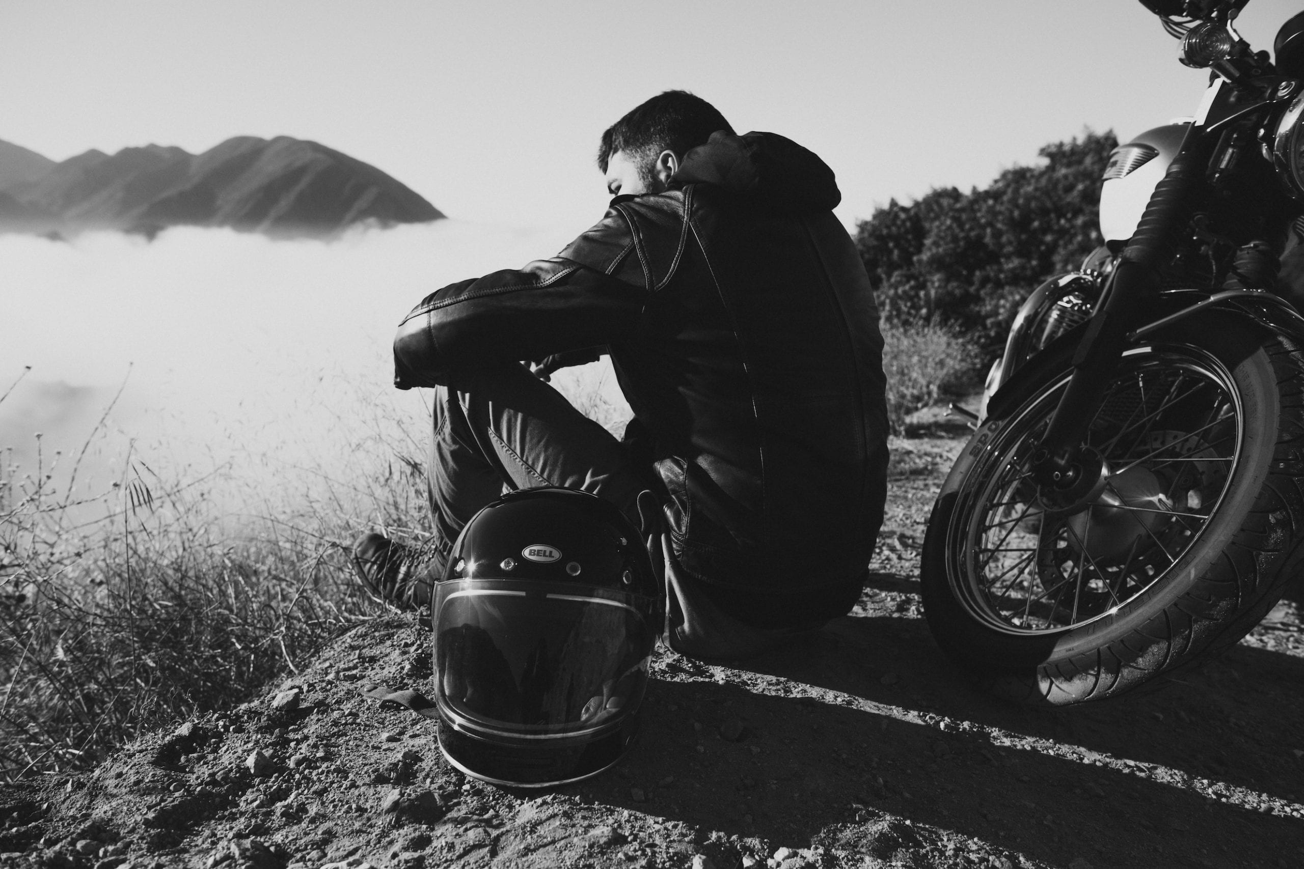 The motorcycle rider relaxes on the side of the road with his Bell Bullet helmet and motorcycle nearby.