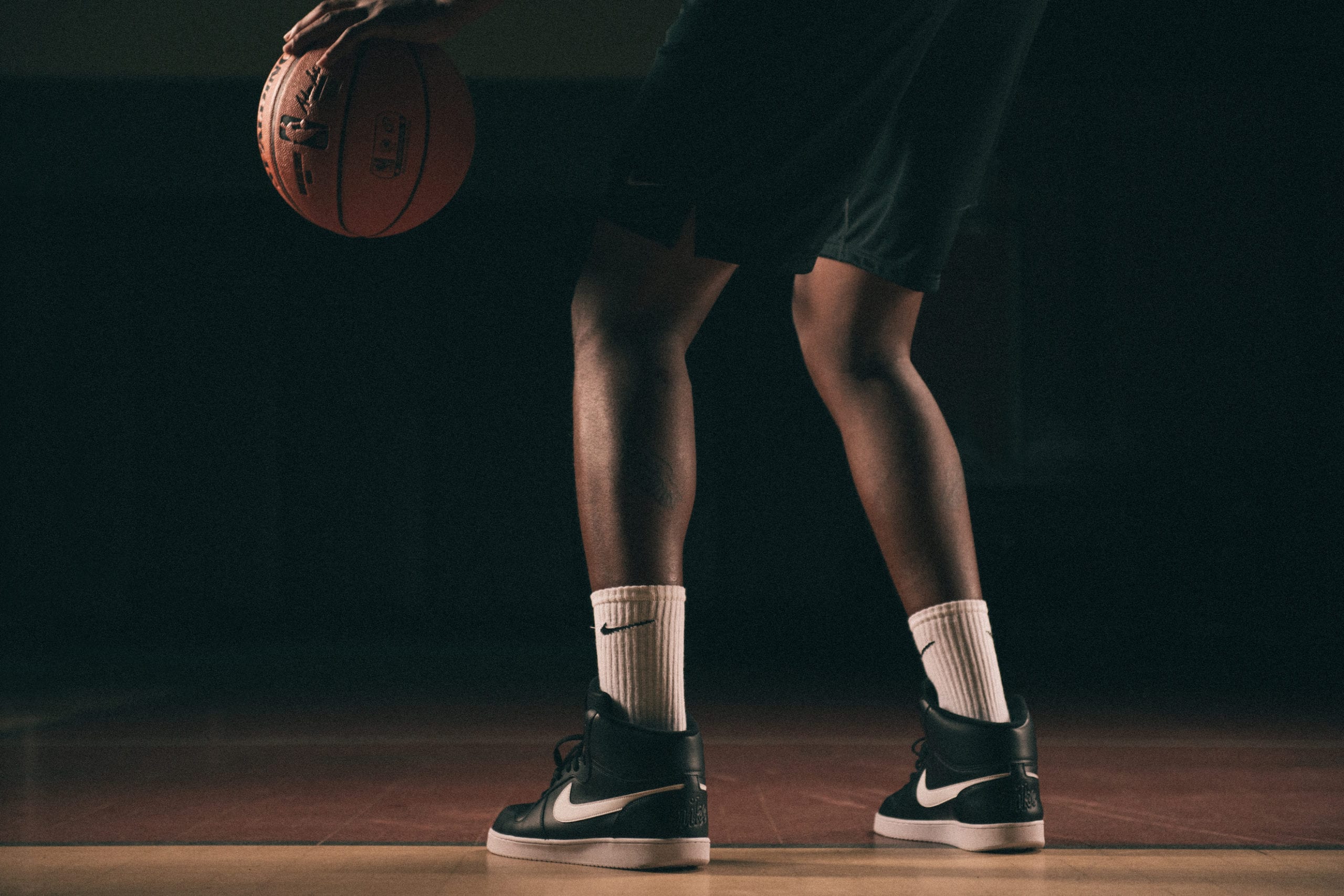 View from behind of the lower half of an African American man wearing black Nike shoes and white Nike socks poised holding a basketball on a basketball court with a light shining on him in semi darkness