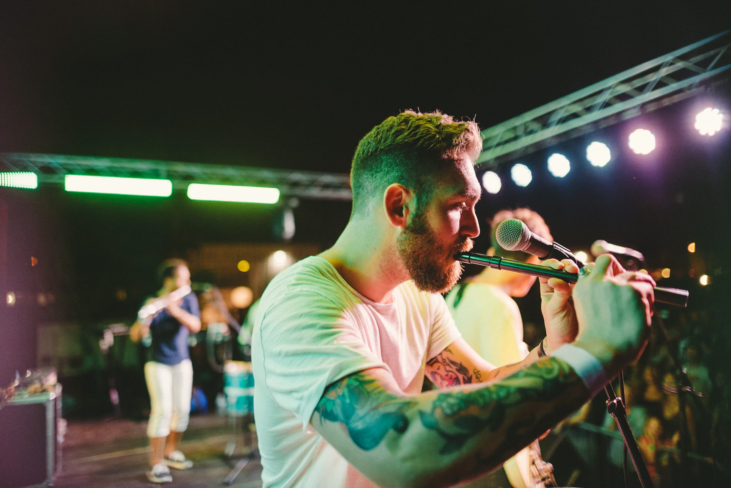 Tattooed short haired with beard flute player playing the flute in a band with another flute player in the background and a guitarist next to him