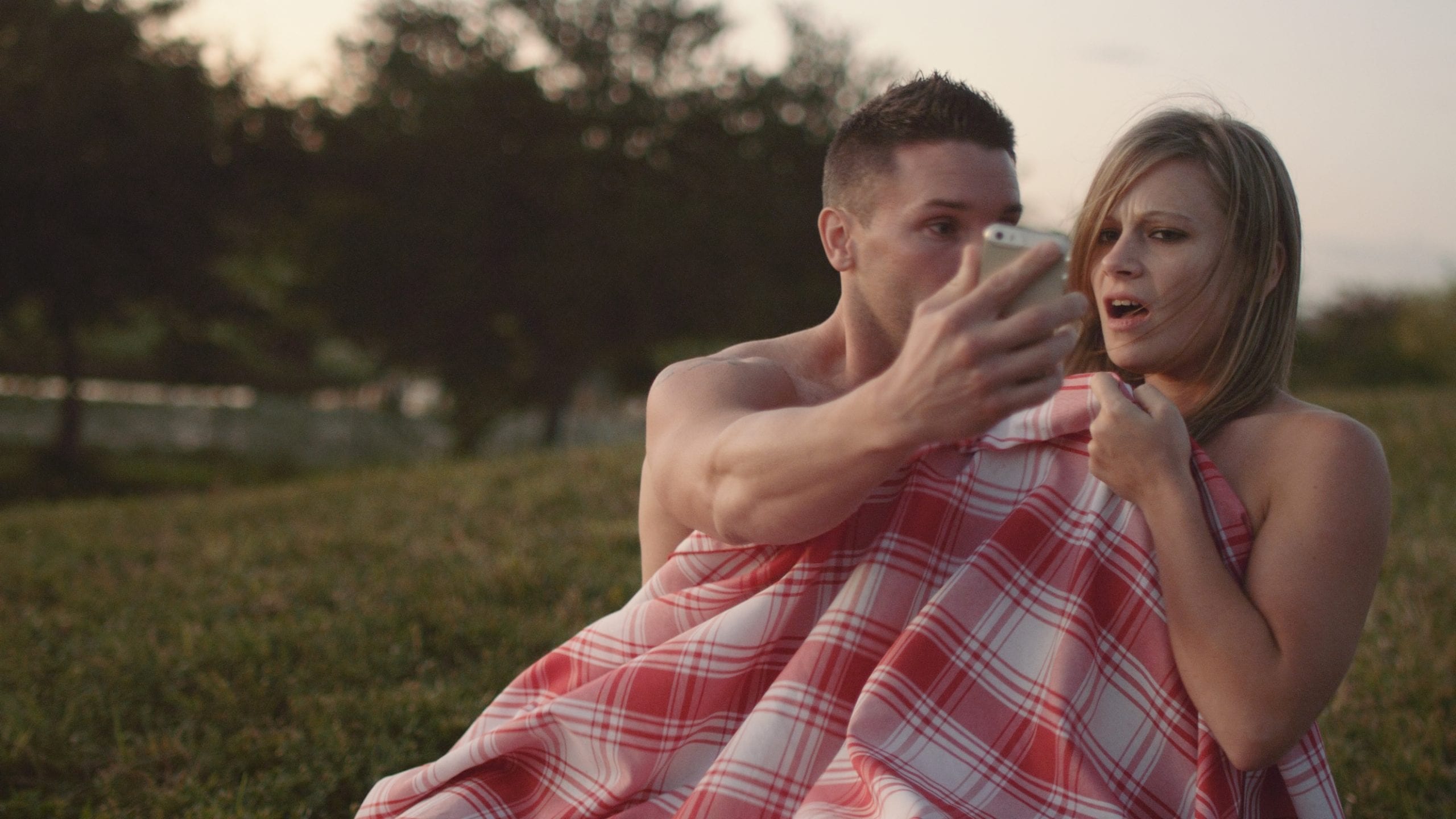Young man and woman under a red and white blanket with man showing woman something on his phone and woman reacting negatively to it