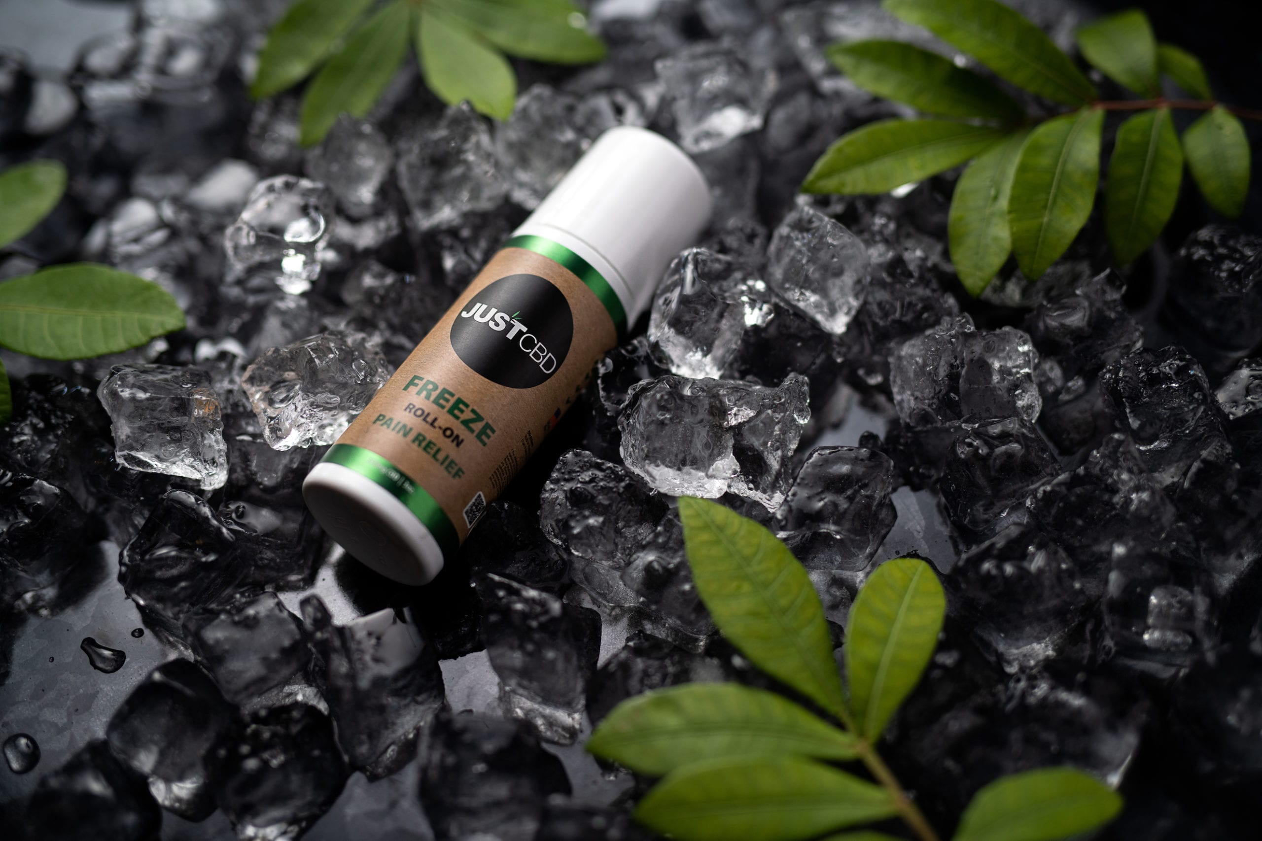 JustCBD Product Photography and Just CBD Products Freeze roll pain relief container on display on a pile of ice with green plant leaves nearby