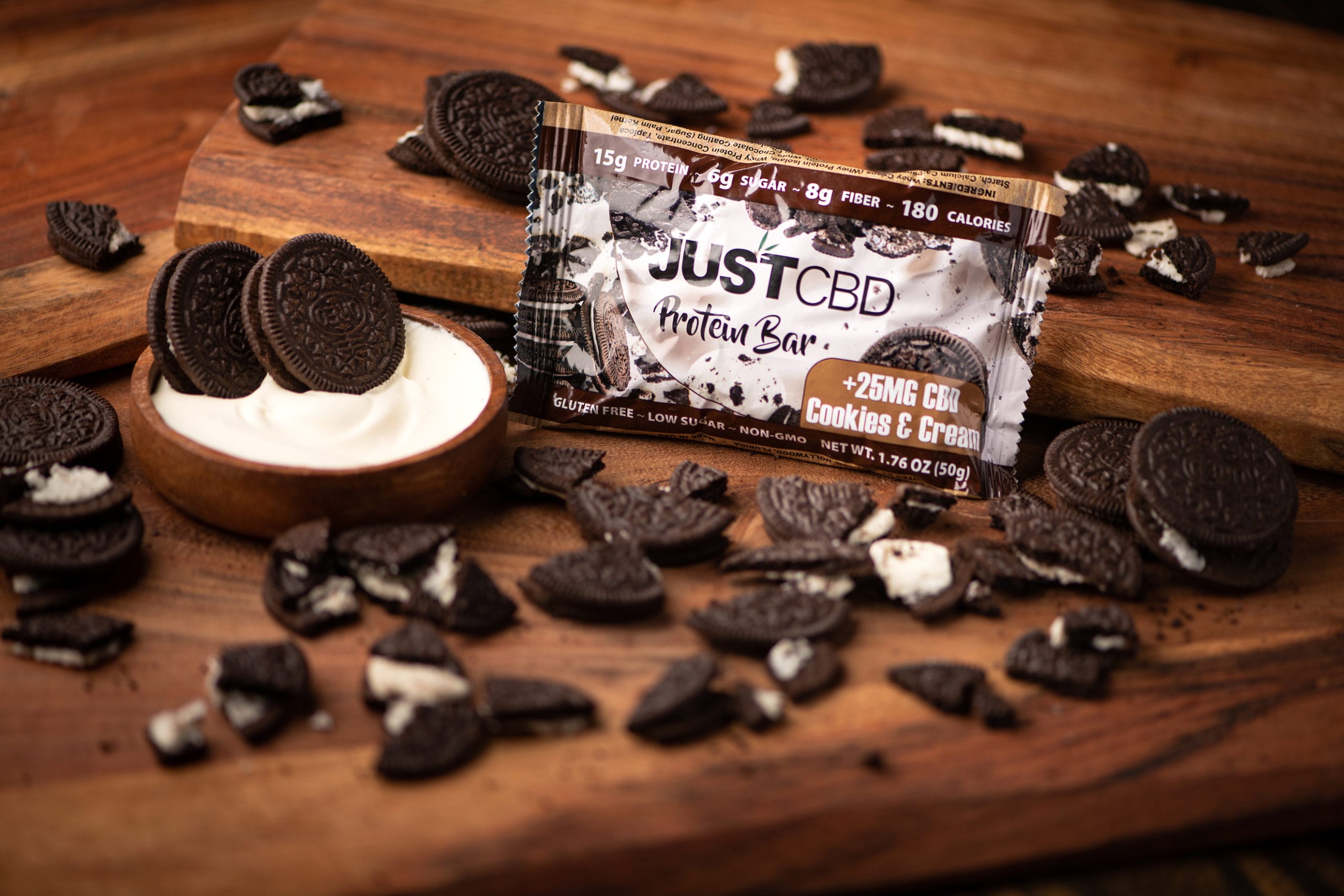 JustCBD Product Photography and Just CBD Products JUSTCBD Cookies and cream protein bar package surrounded by cookies and bowl of cream on a wooden table