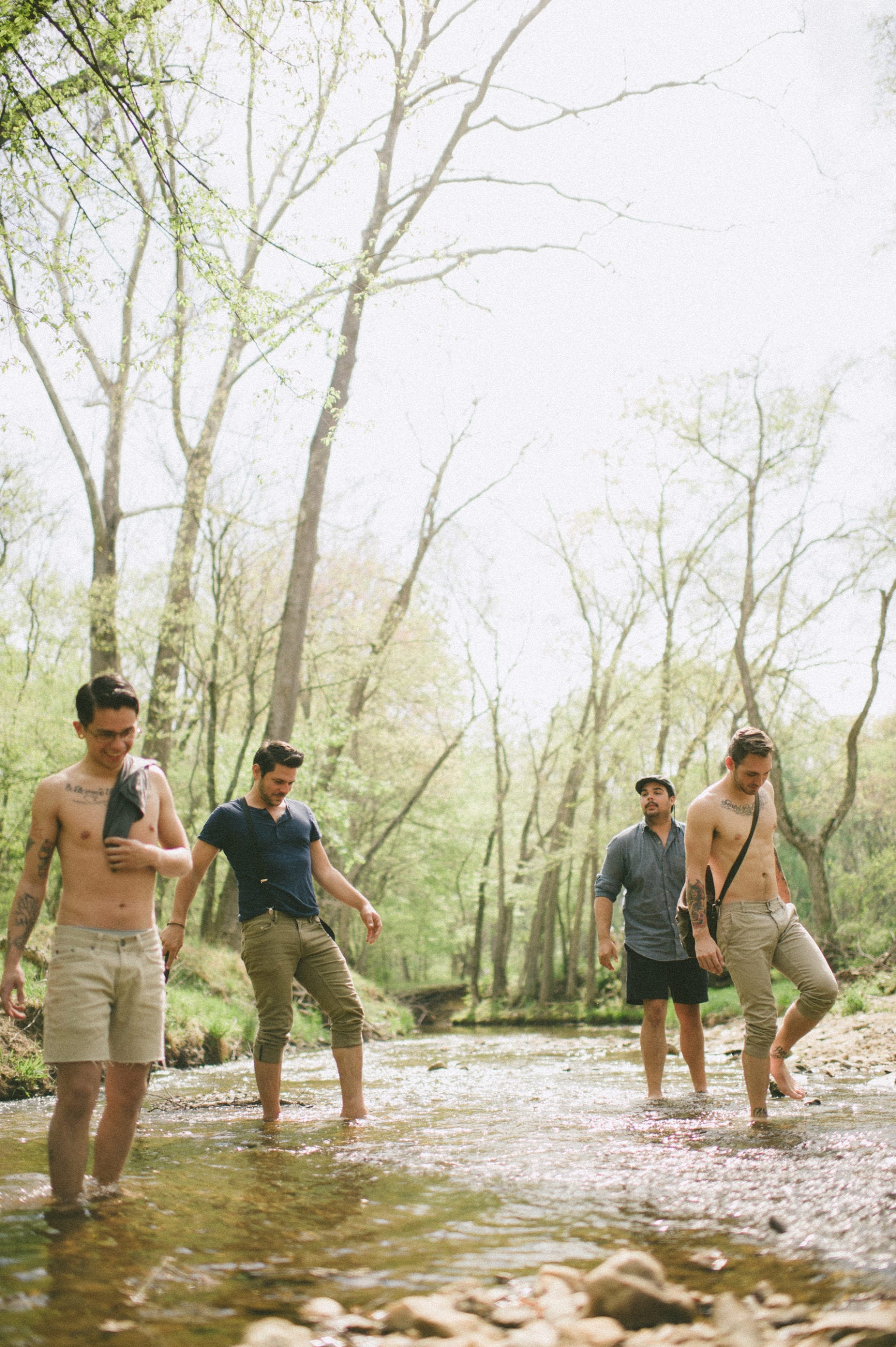 Four males walking in a creek on a sunny day in the forest. Two males are shirtless with tattoos on their arms and chest.