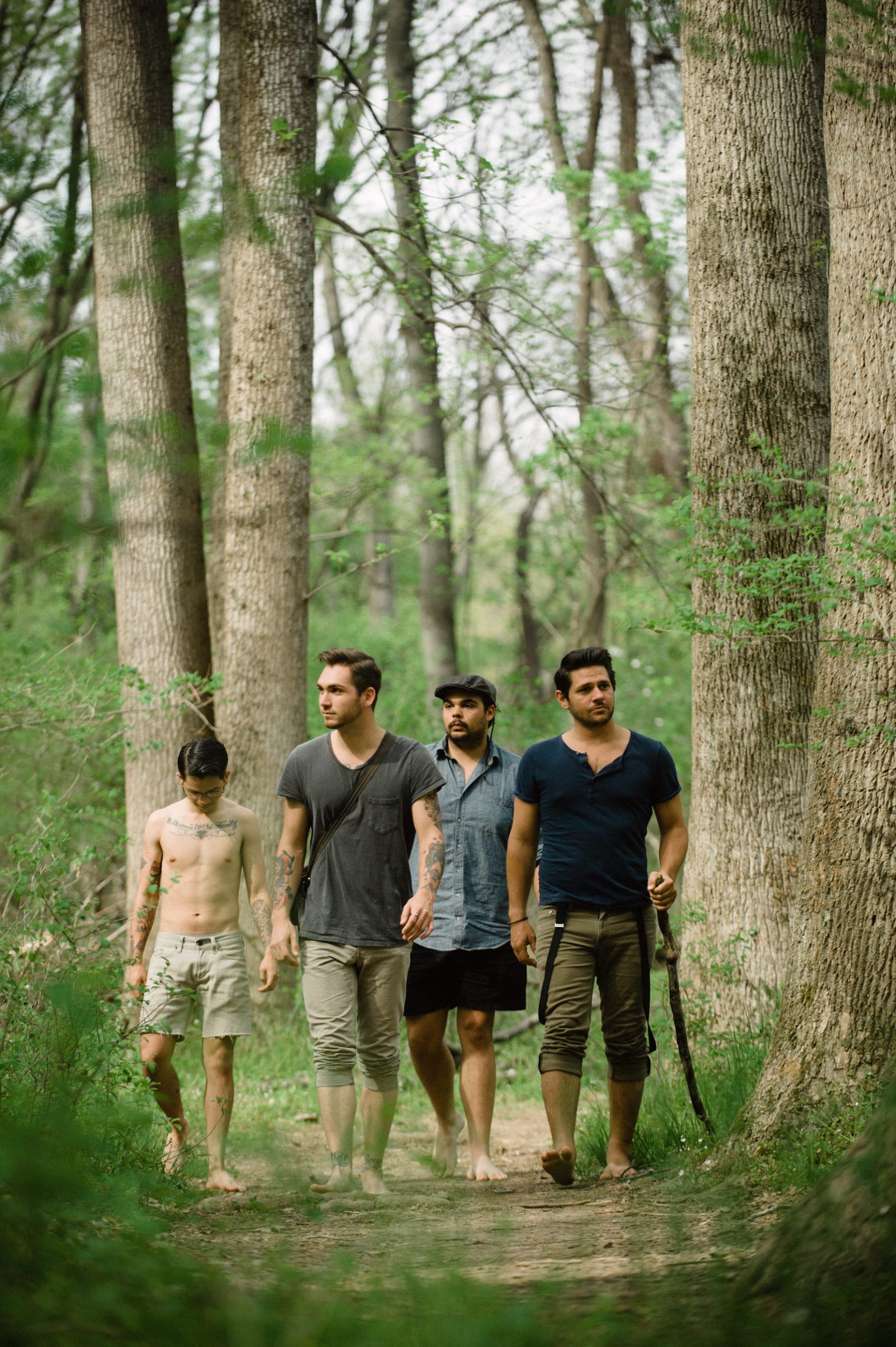 Four men walking in a forest with one shirtless with tattoos and one holding a stick