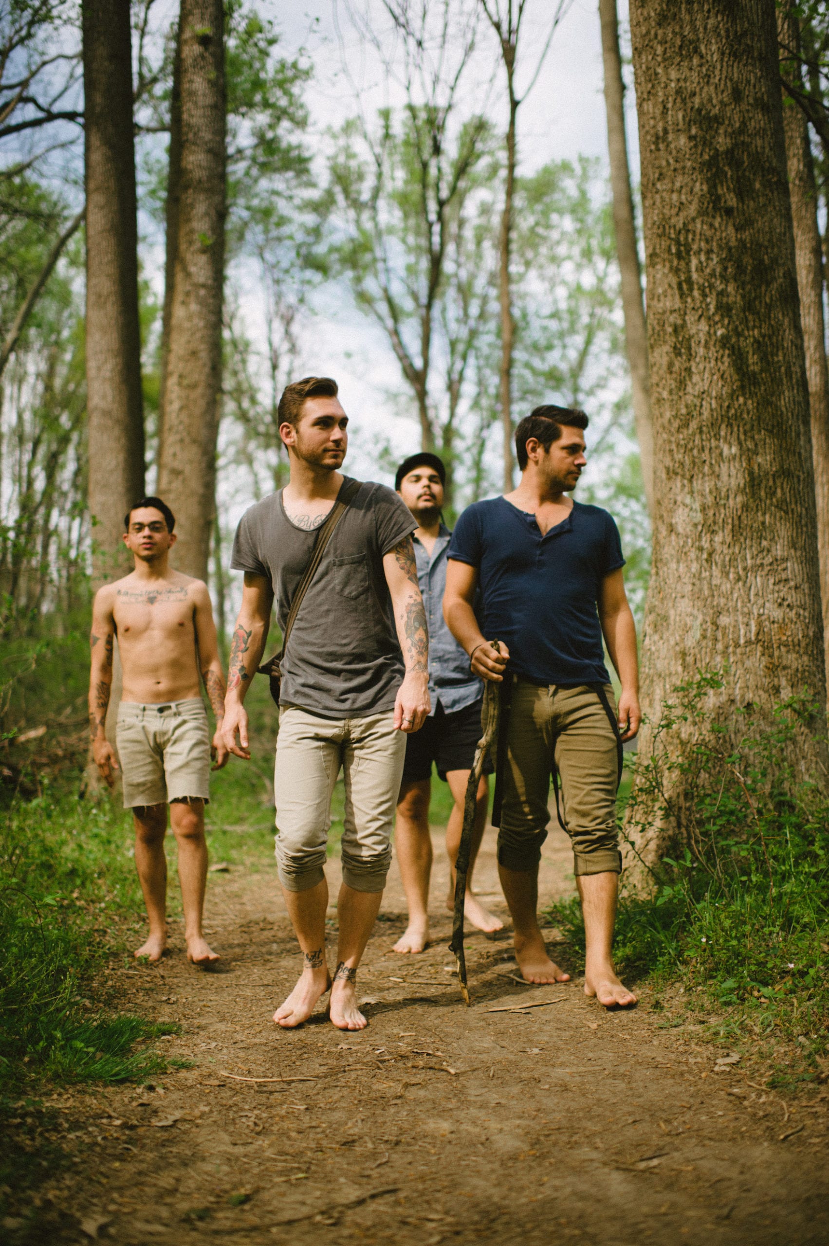 Four males walking in the forest on a sunny day. One is shirtless with tattoos on arms and chest. Two males are looking ahead into the distance while the other two are looking to their left.