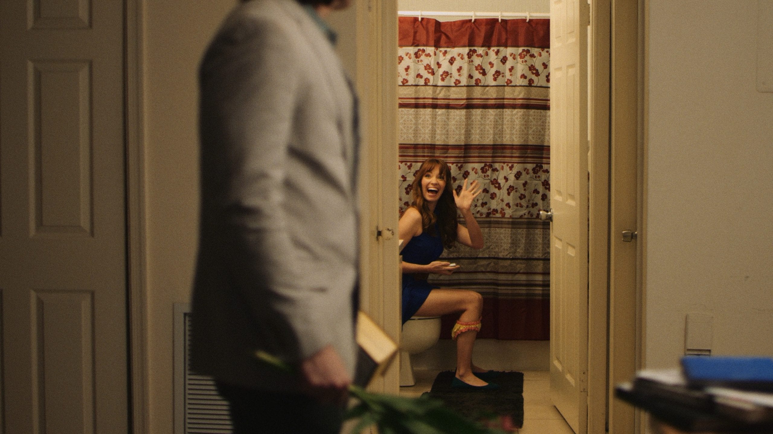 Man in a gray suit carrying a box of Whitman's Chocolates and flowers walking past a bathroom with a woman sitting on the toilet waving and smiling