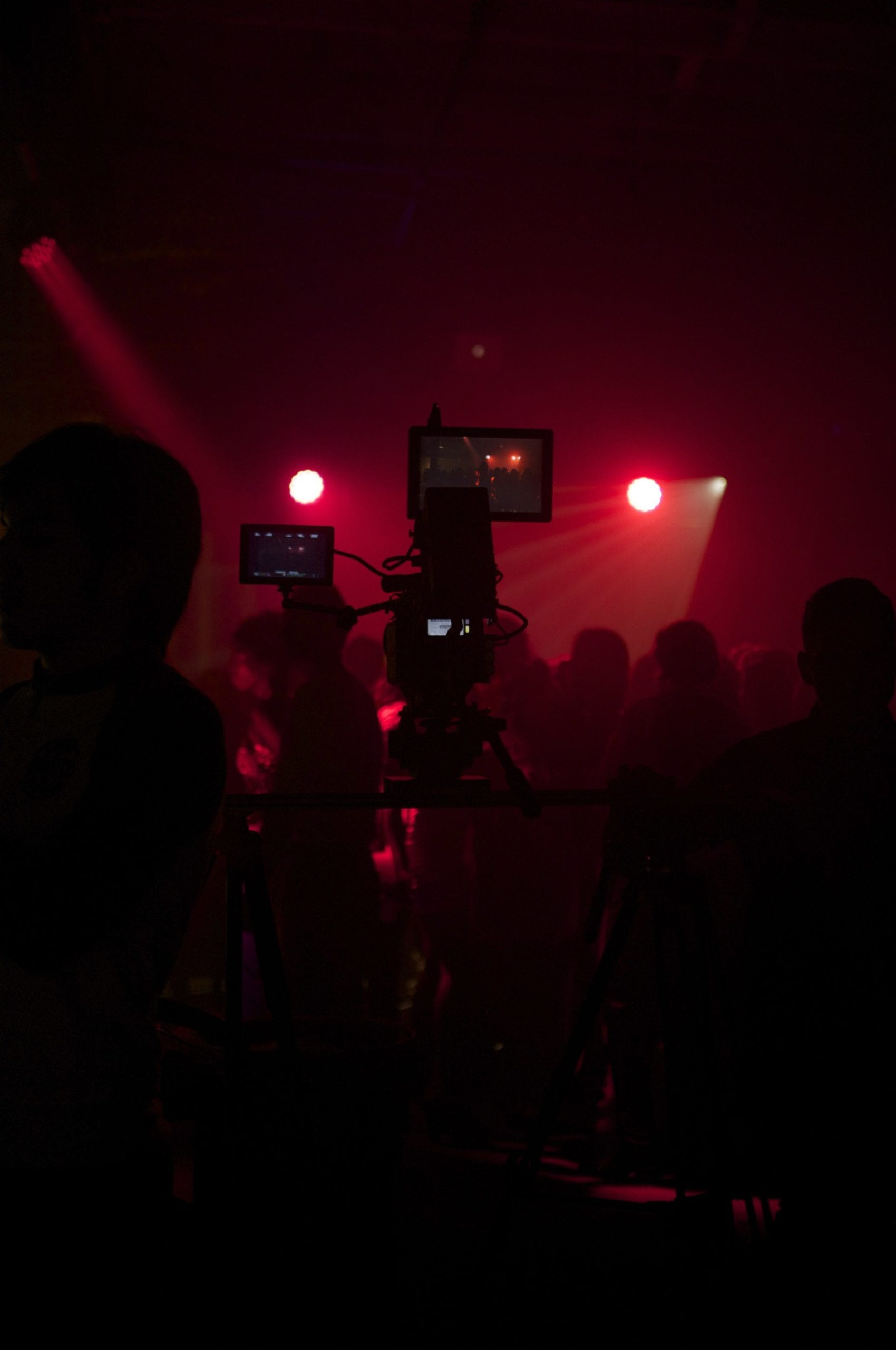 View from behind silhouette of two video camera operators filming a concert basked in red light