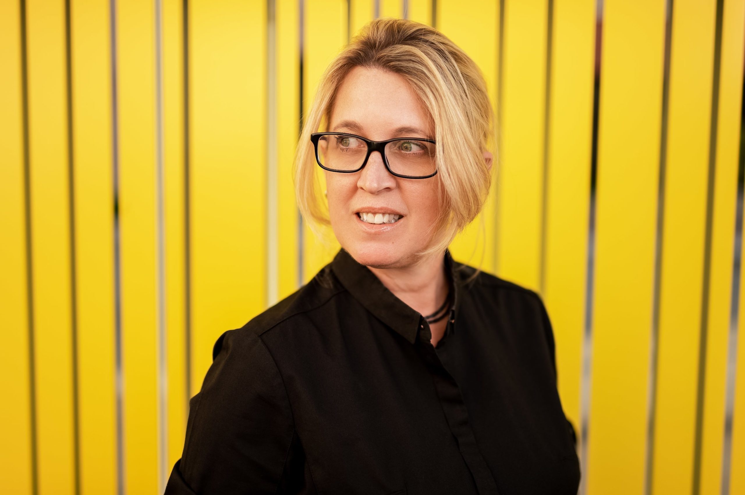 Glavovic Studio Fort Lauderdale Headshot of woman with short blond hair and glasses wearing a black dress shirt posing for camera in front of yellow wall looking off to the side