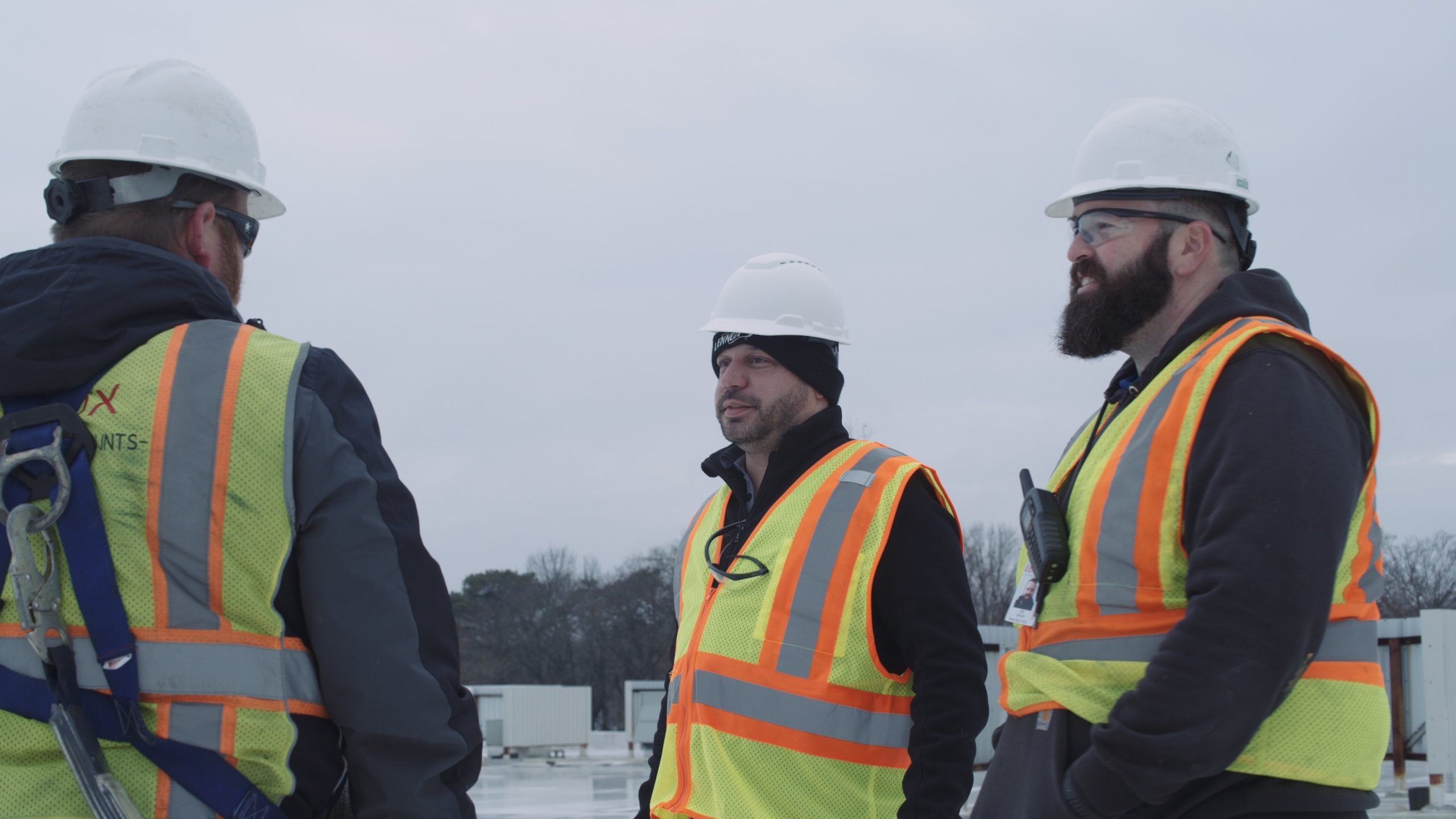 Three workers with beards wearing reflective safety vests and white hard hats talking to each other