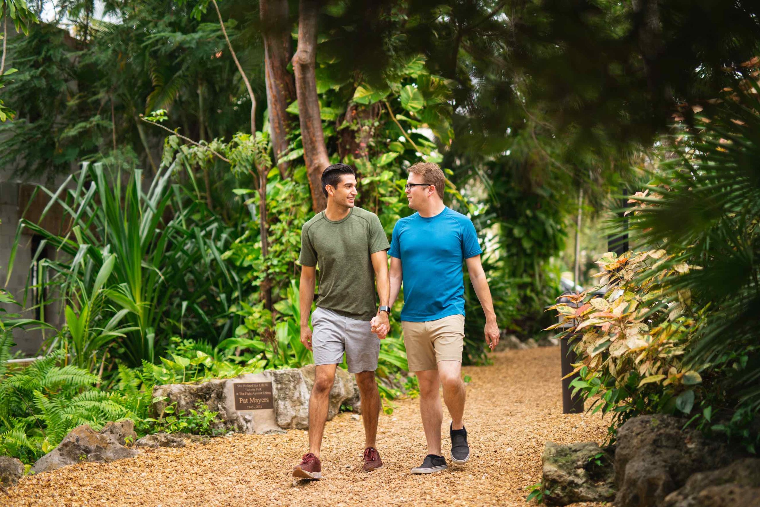 Two men both wearing t shirts and shorts, holding hands and walking down a path in the woods looking at each other